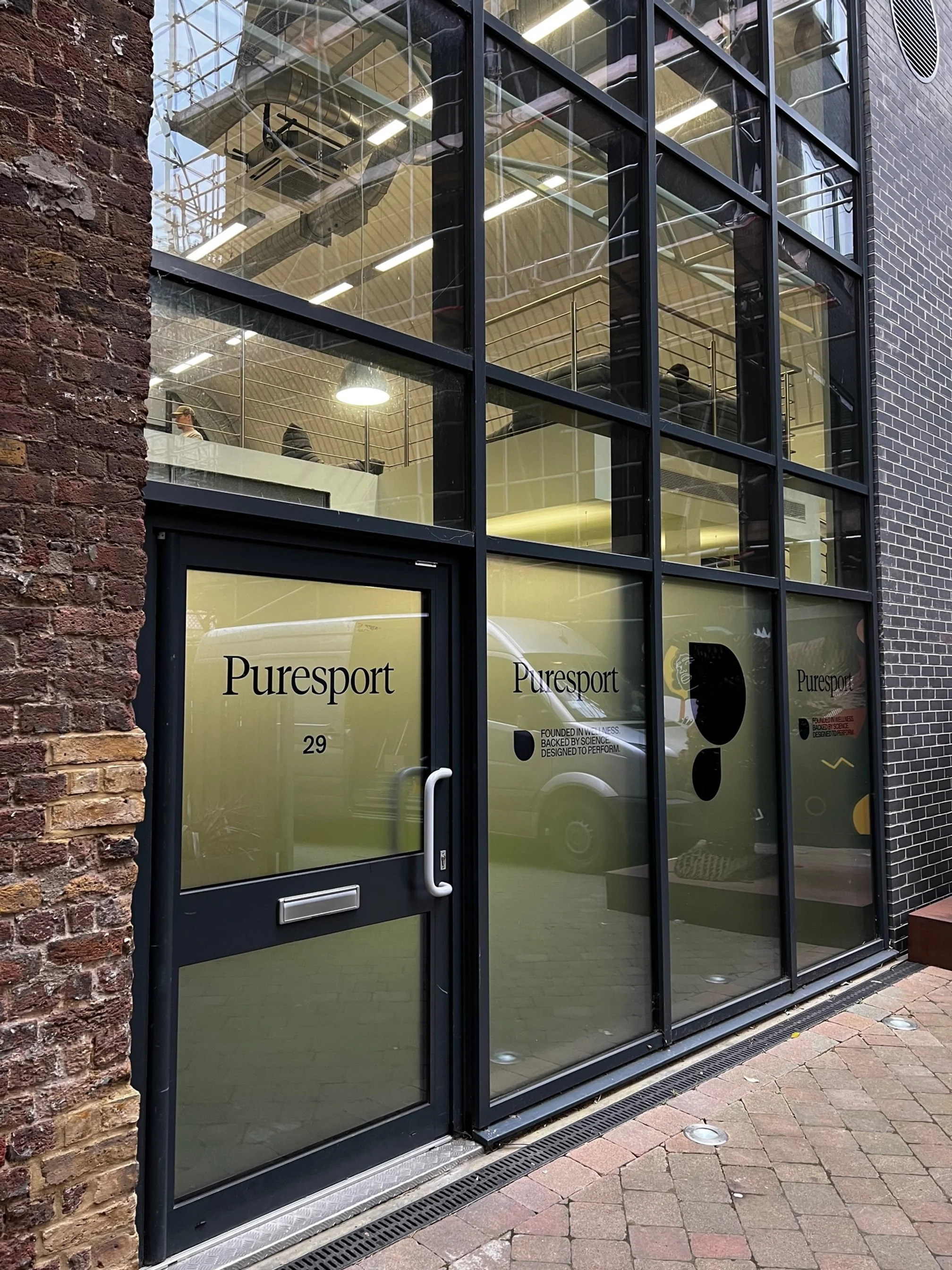 Glass storefront of Puresport with a door, showing the company name and logo, part of a brick building, with an interior visible through the windows and people inside.