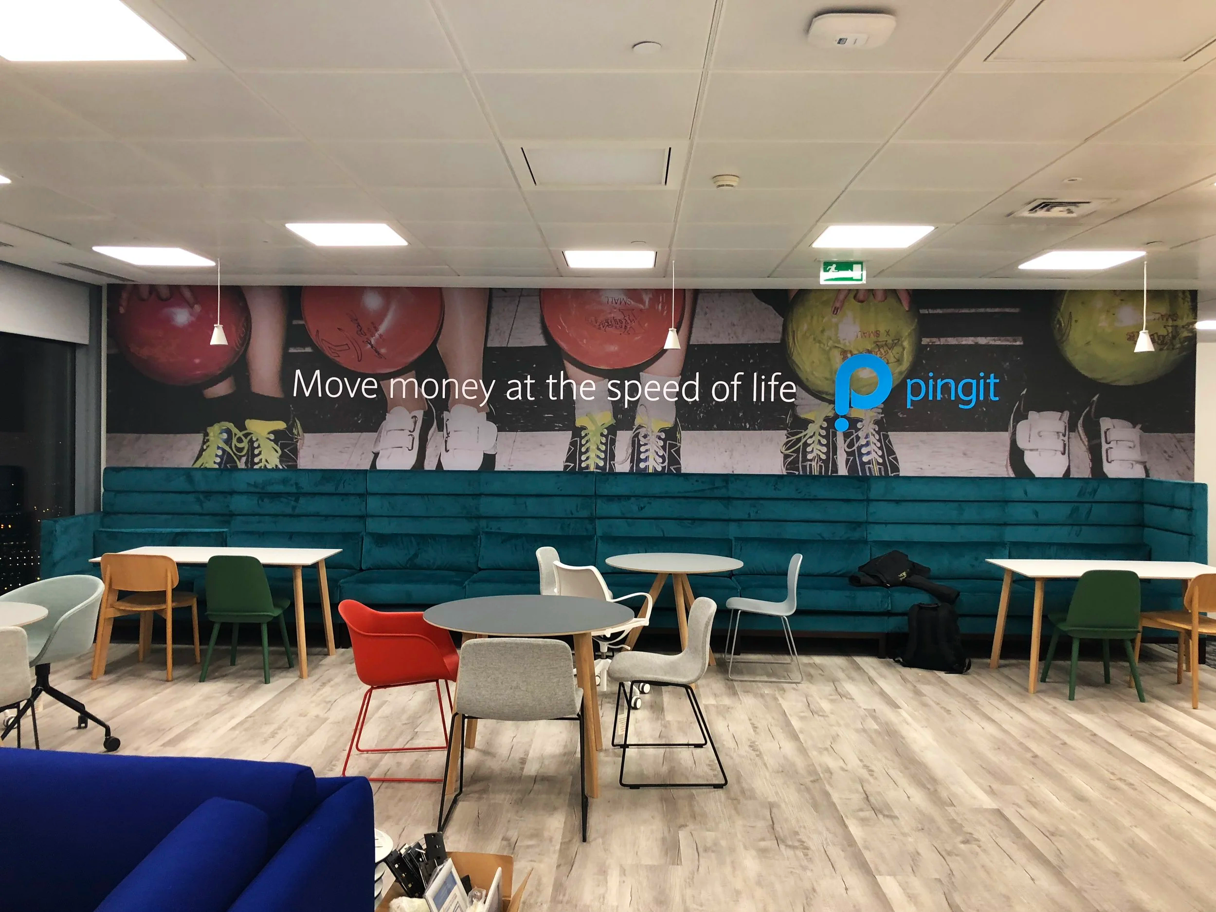 Interior of a modern social space with colorful chairs and a teal couch, a large mural of people holding bowling balls with a message about moving money quickly, and a Pingit advertisement.