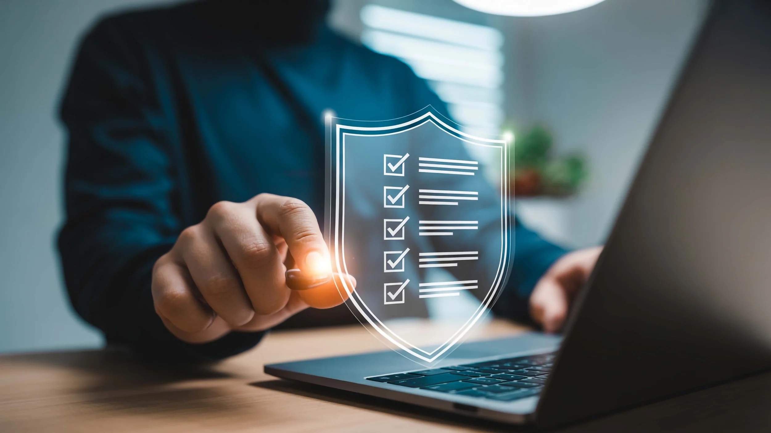 Person in a dark shirt using a laptop with a virtual shield with checklist icons projected in front of them, symbolizing cybersecurity or online protection.