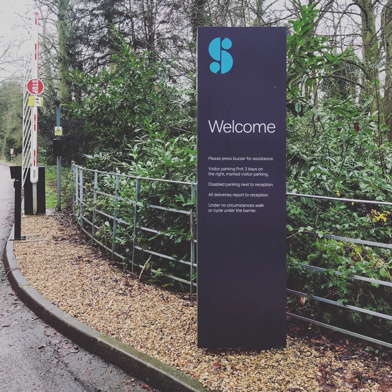 A large blue information sign with a logo and welcome message next to a wooded area, with instructions for visitors and parking information.