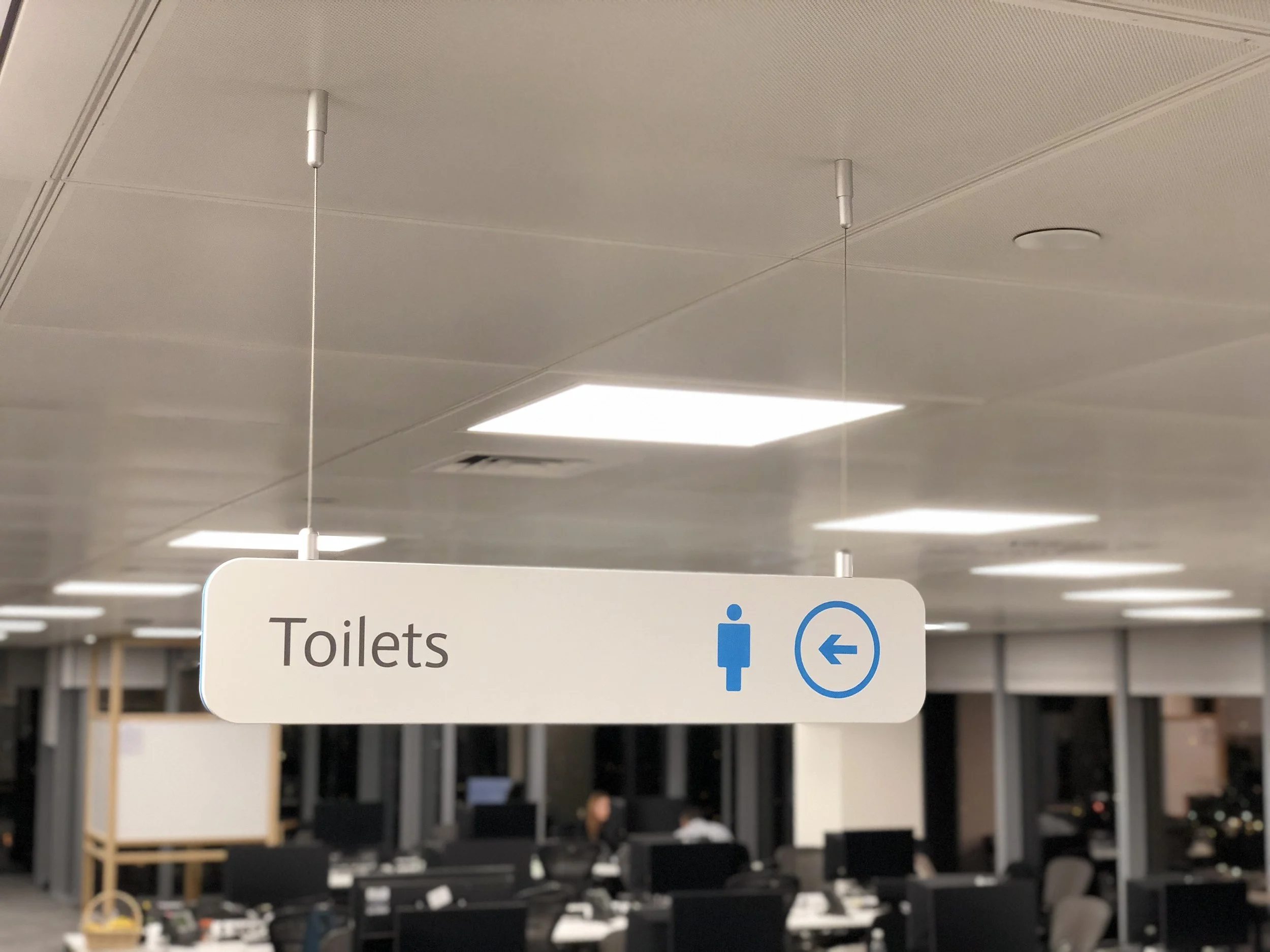 A directional sign indicating the location of toilets, with symbols for male and female, in an indoor setting, possibly an office or airport.