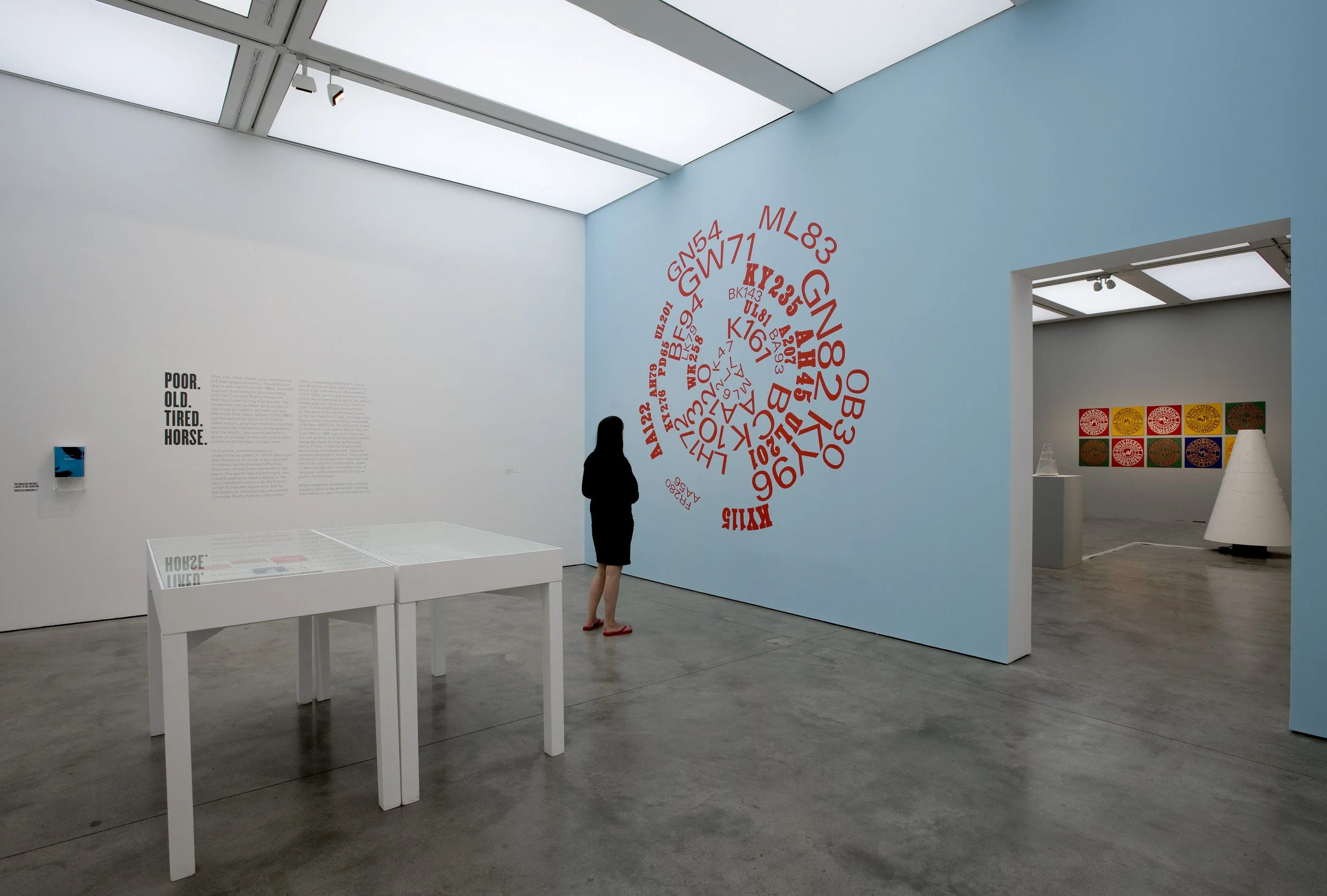 An art gallery with white walls and a blue accent wall displaying a large red spiral of letters and numbers. There is a silhouette of a person in black standing in front of the wall, with a display case on a white table nearby. On the left wall, there is text that reads 'POOR. OLD. TIRED. HORSE.' in black. In the background, another room contains colorful square artworks and cone-shaped sculptures.