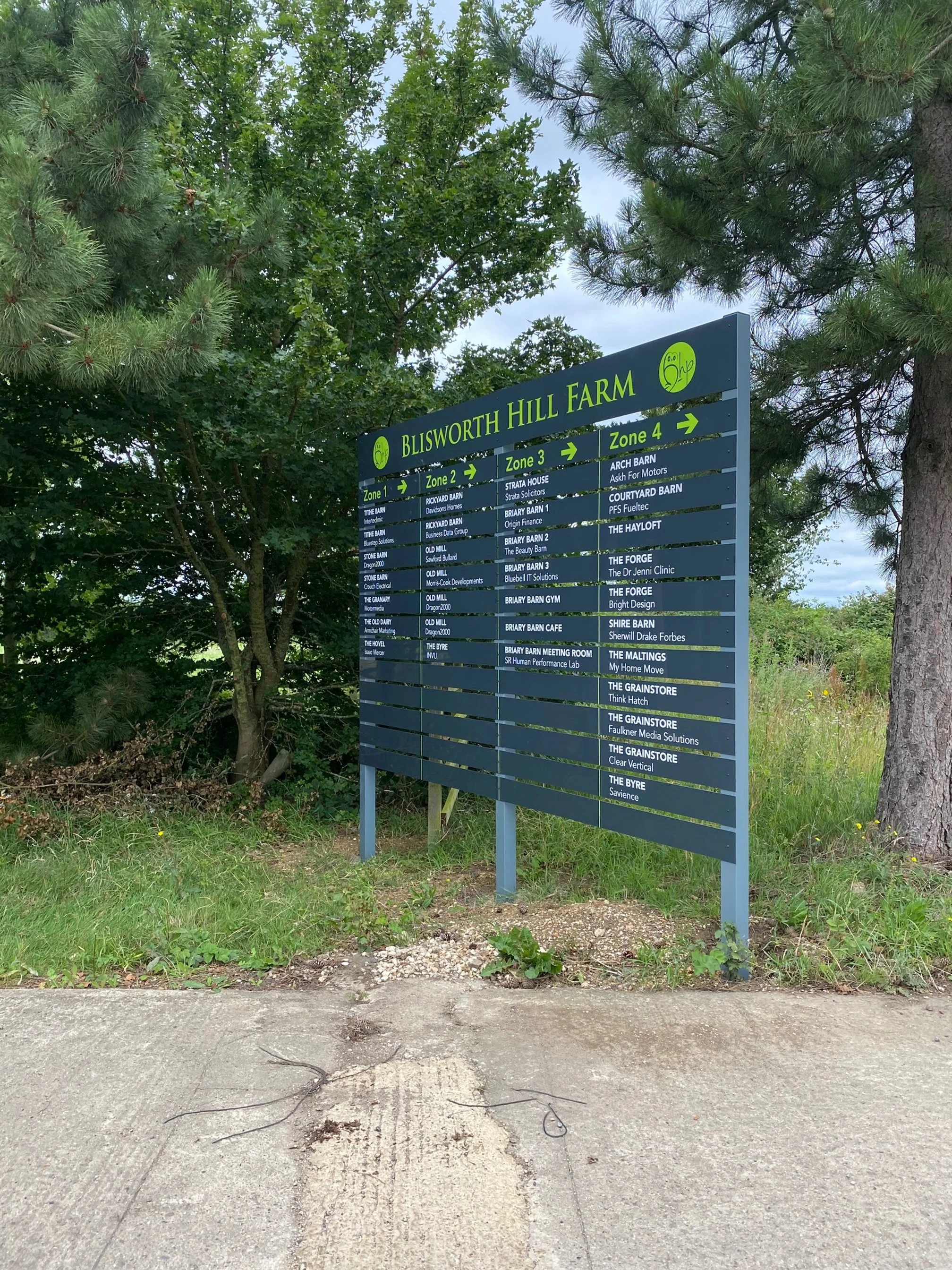 Signboard for Blisworth Hill Farm showing zones and businesses, surrounded by trees and grass.
