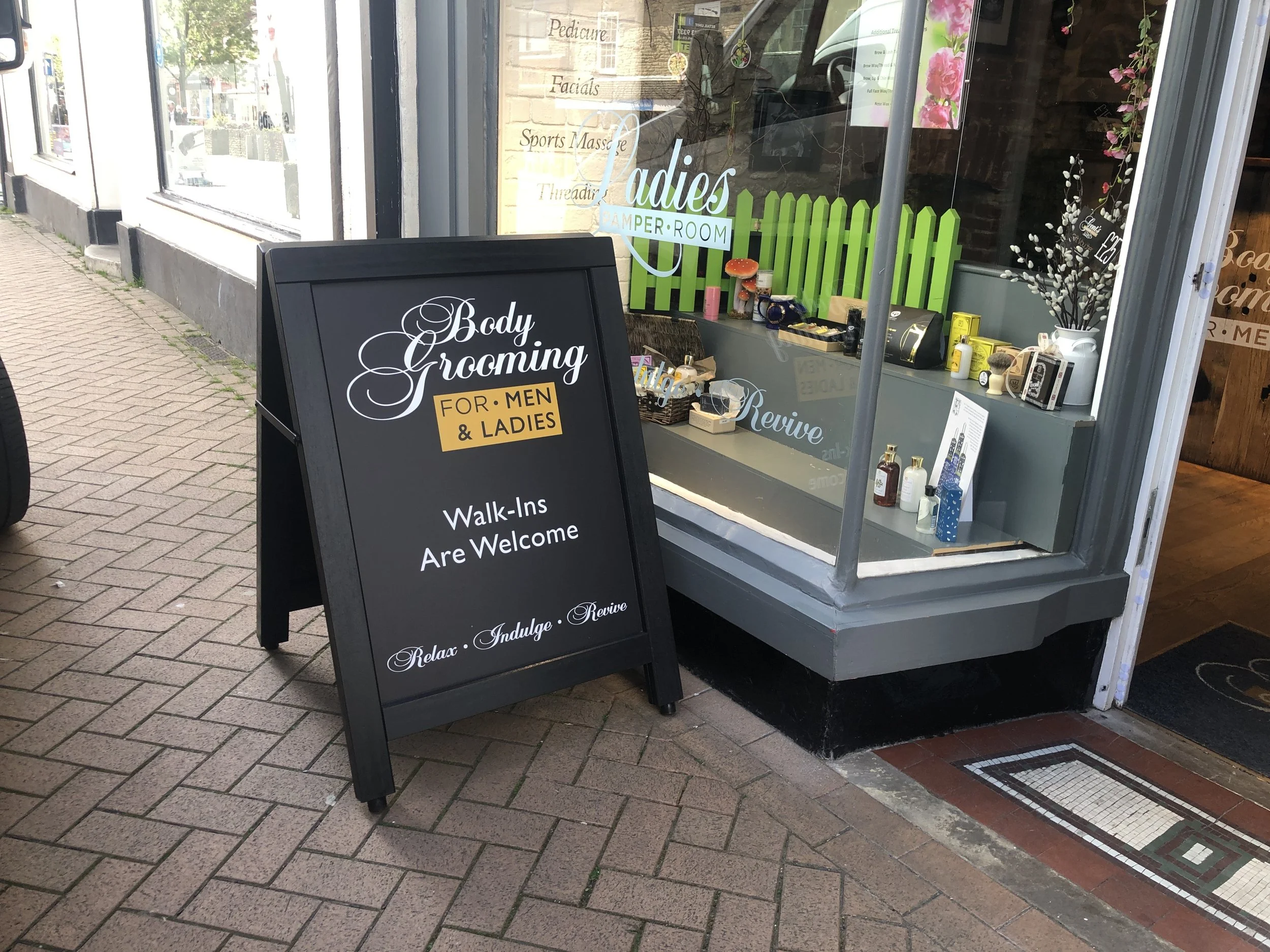 A black sidewalk sign in front of a store advertising body grooming services for men and women, welcoming walk-ins, with phrases: 'Relax. Indulge. Revive.' The store window displays various products, flowers, and a green fence decoration.