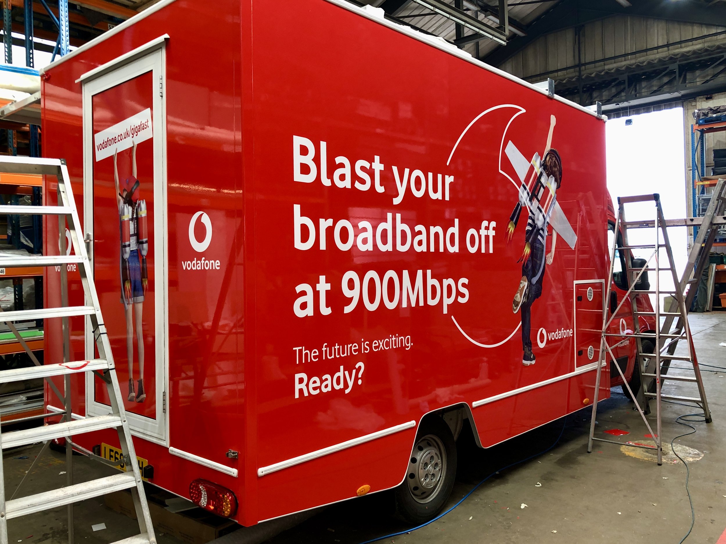 A red mobile advertising truck with a large promotional message for Vodafone, featuring an animated character with rocket-powered shoes on the side. The truck is inside a warehouse with ladders nearby.