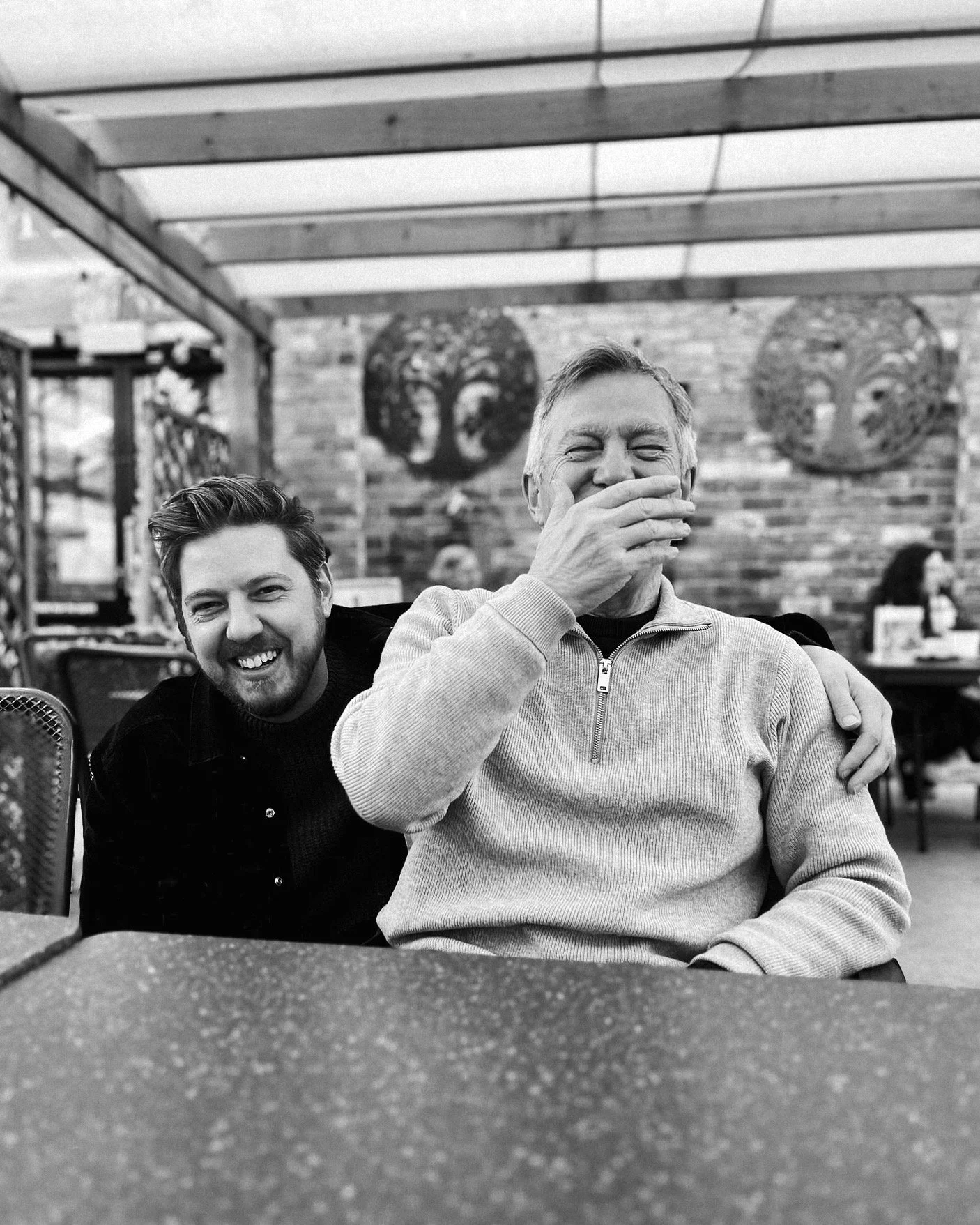 Two men sitting at a table in a restaurant or cafe, smiling and laughing, with one man covering his mouth with his hand. The background has brick walls and artwork.