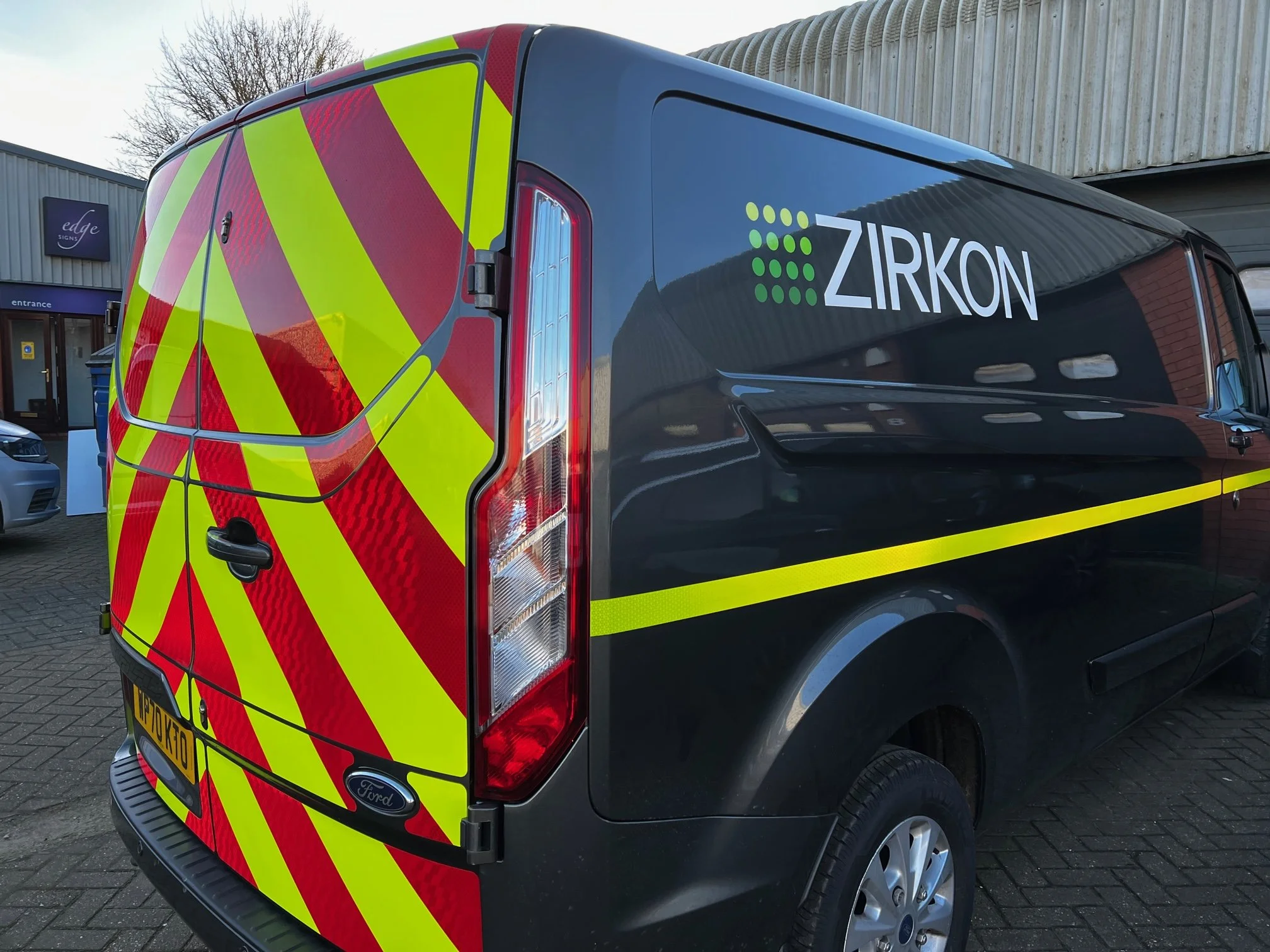 A black Ford van with yellow and red reflective safety markings parked outside a building. The van has the company name ZIRKON on the side and is parked on a brick driveway.