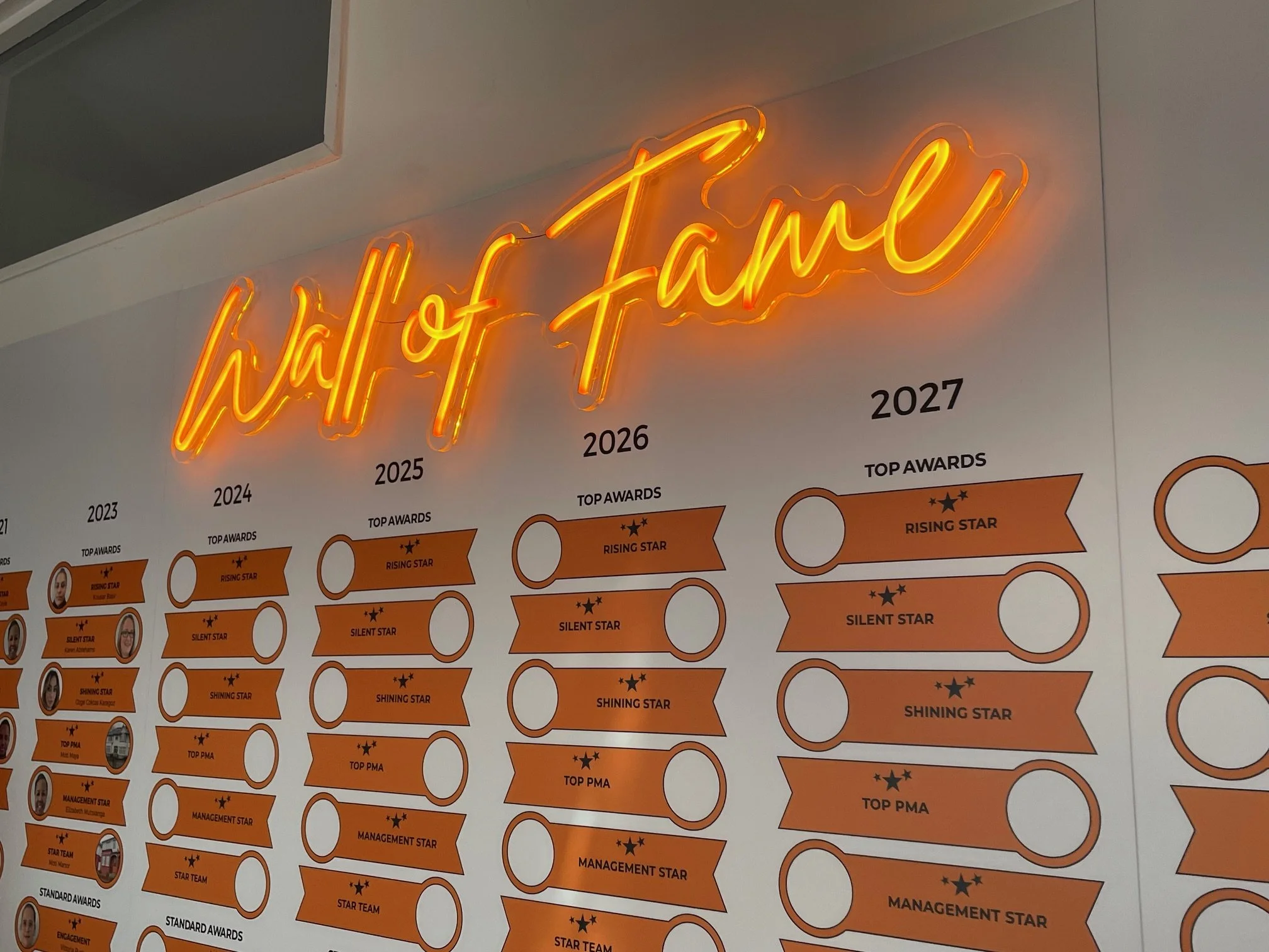 A wall display with a neon sign that reads 'hall of fame' in orange cursive, and a timeline from 2023 to 2027 showing various awards and achievements for each year.