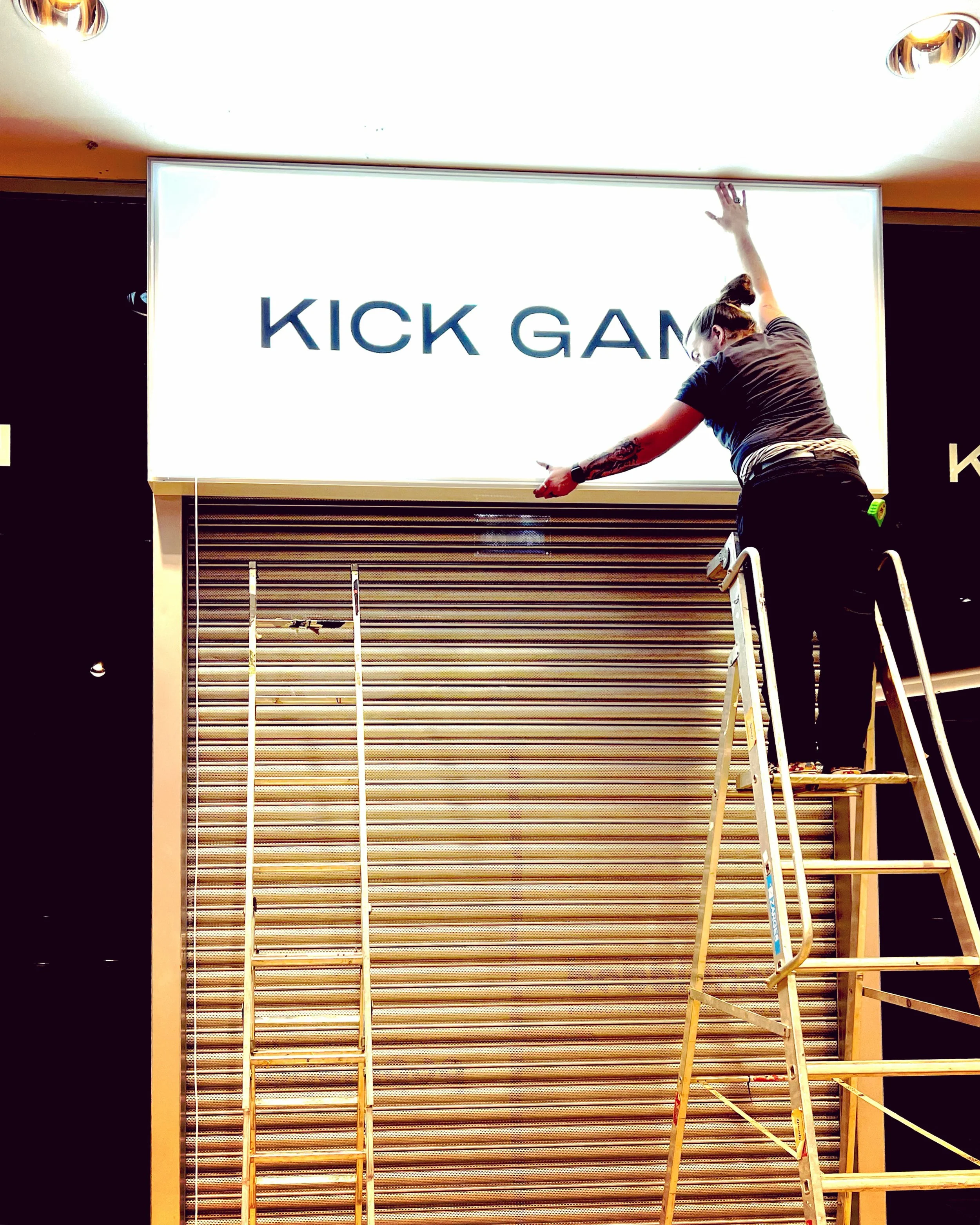 A worker on a ladder arranging a backlit sign that reads 'KICK GAM' in a retail store.