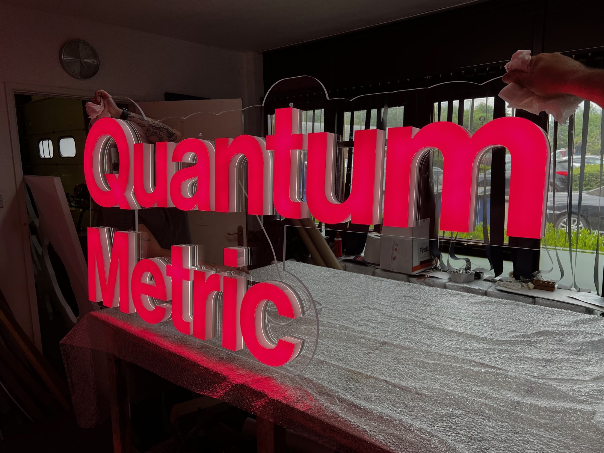 Red illuminated sign that reads 'Quantum Metric' being installed or assembled indoors near a window.