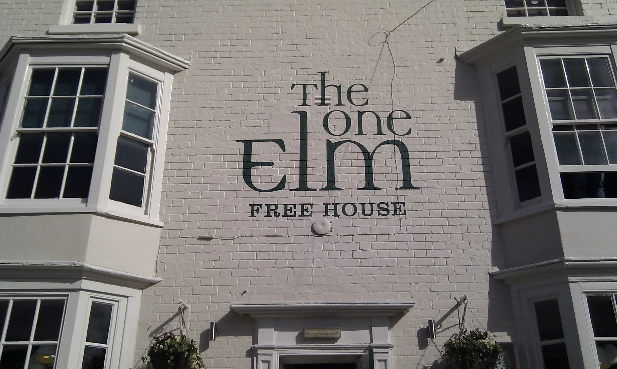 Photo of a white brick building with large windowed bay windows on each side. On the wall, there is a sign that reads 'The One Elm FREE HOUSE'.
