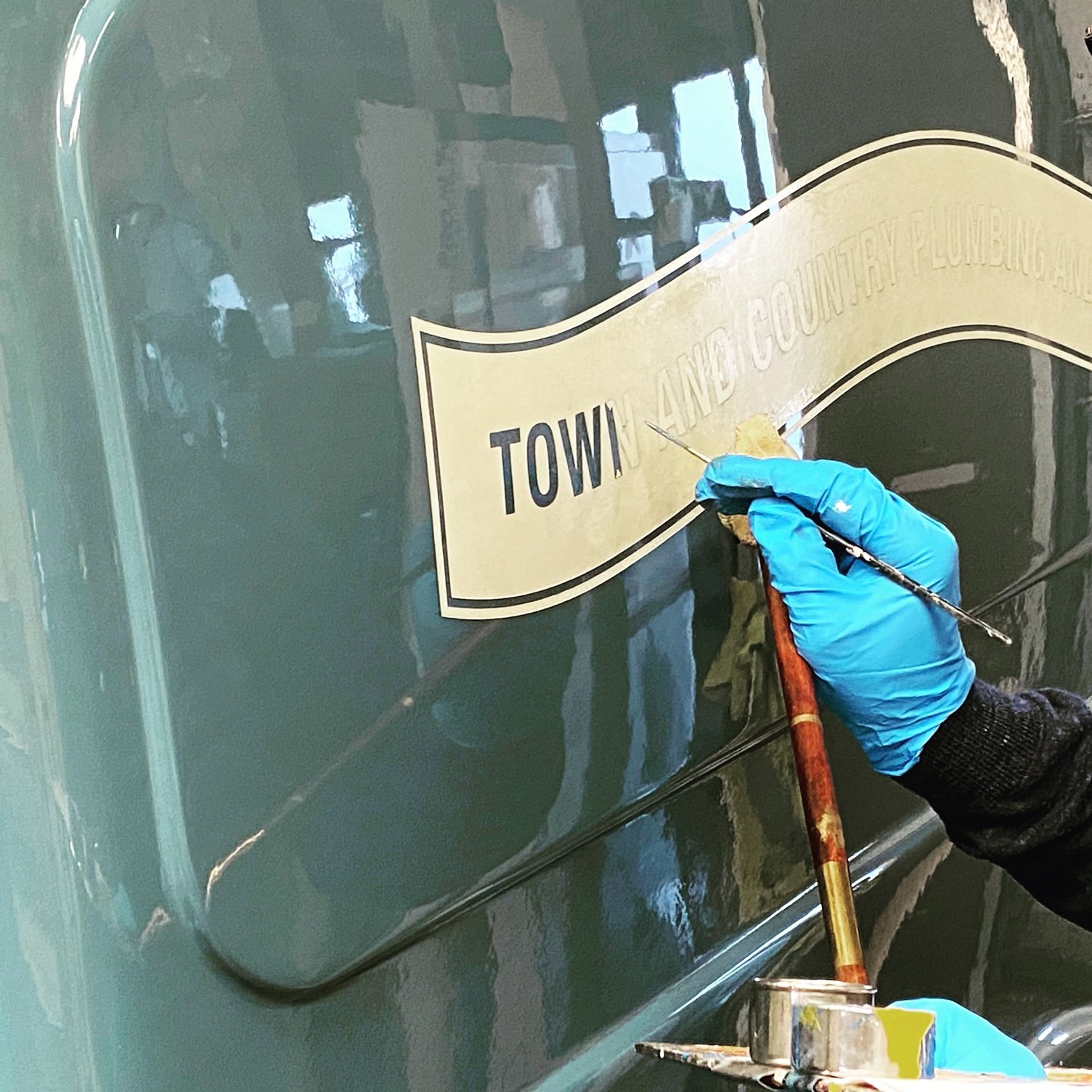 A person wearing a blue glove is painting a sign on a dark green wall with a small brush, the sign reads 'TOWN AND COUNTY PLUMBING'.