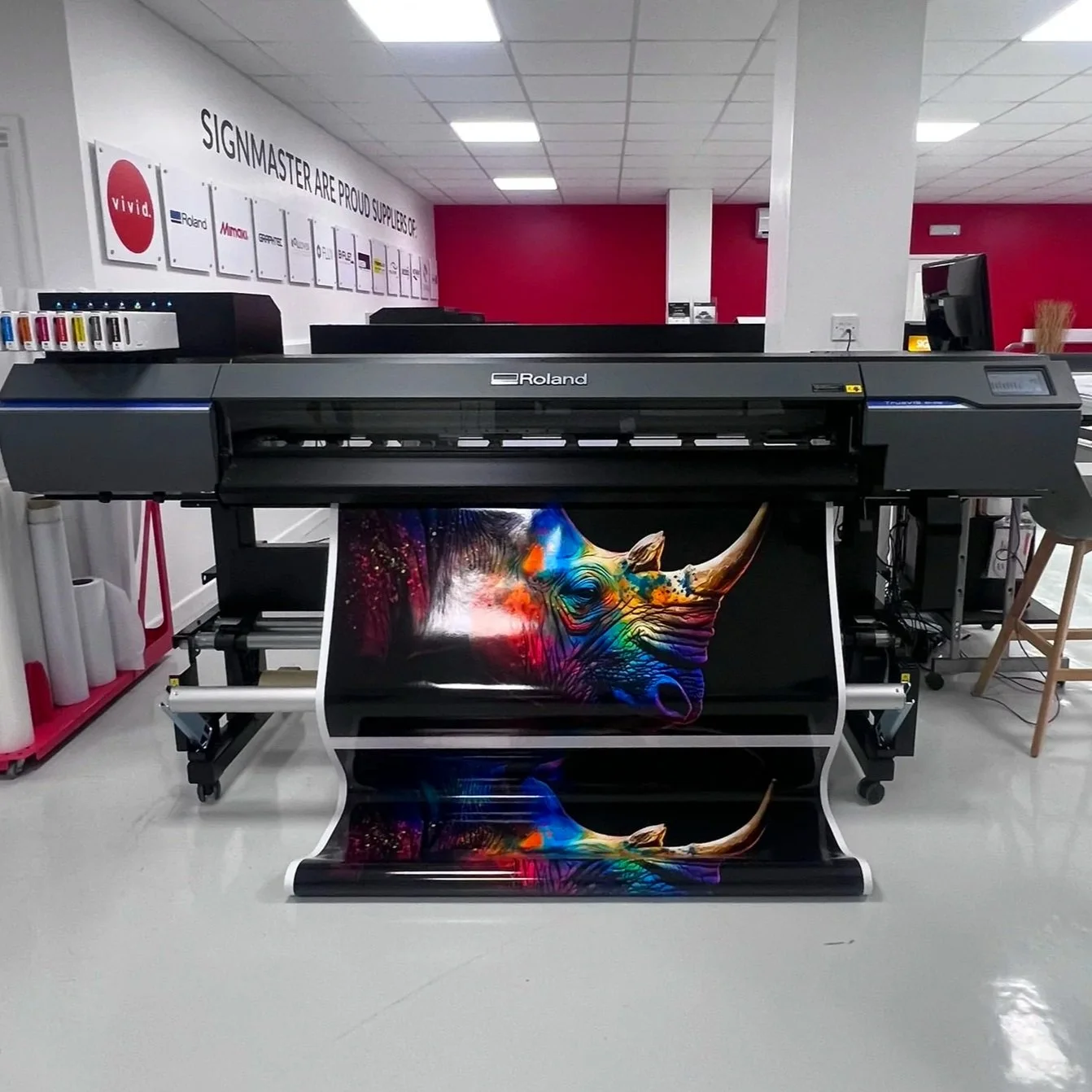 Large Roland printer printing a vibrant, colorful image of a rhinoceros's head on glossy paper in a print shop.