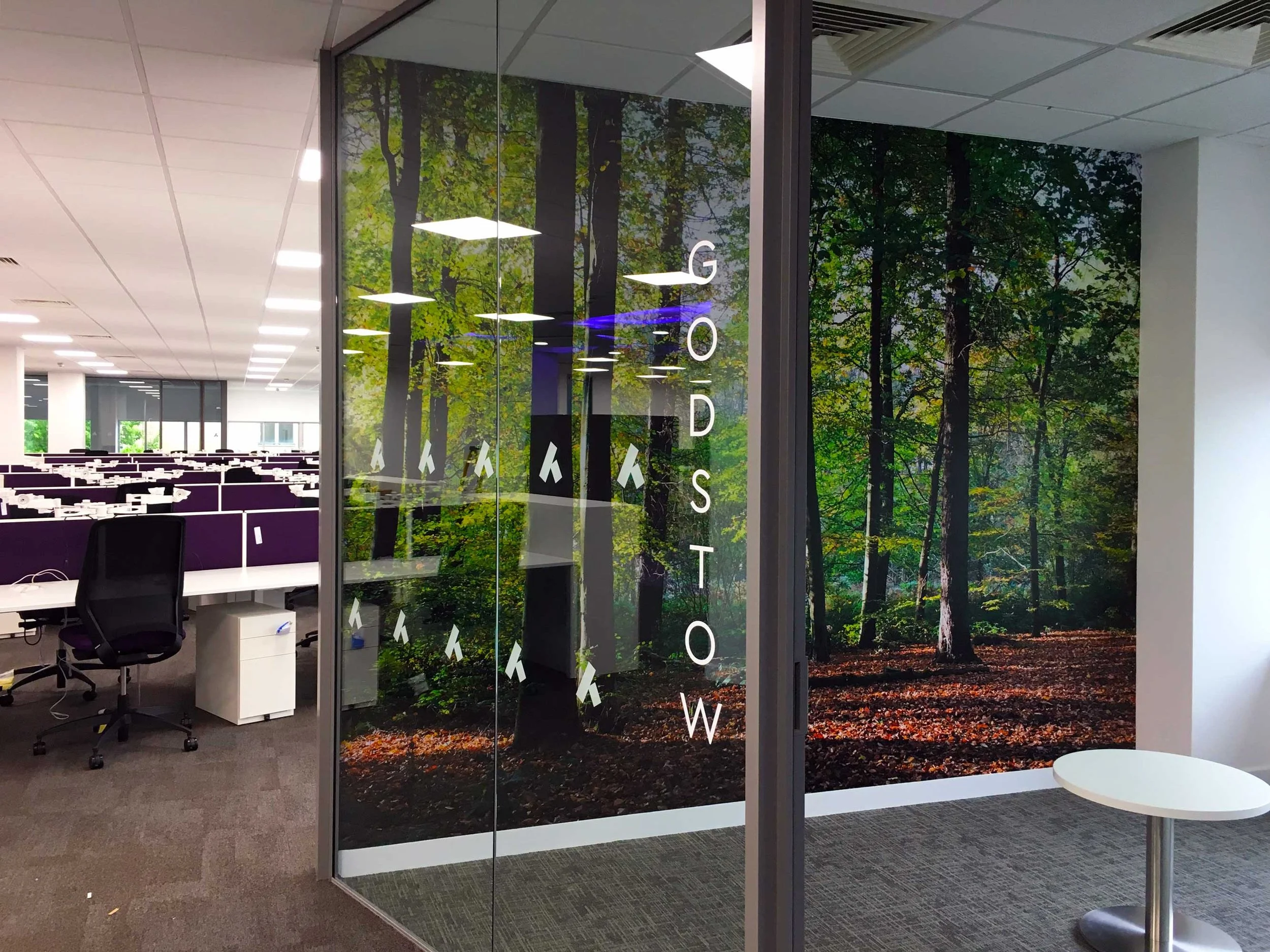 Office space with a glass wall featuring a forest scene and the phrase "GOD'S TOW" written vertically. Inside, there are desks with purple partitions and office chairs, and on the outside, a small white round table.