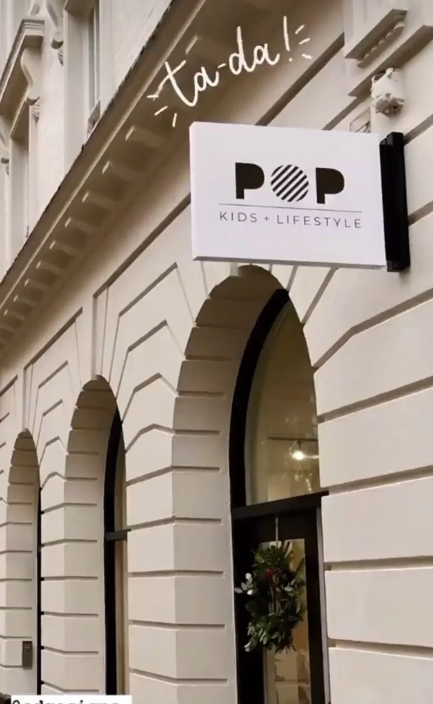 Exterior view of a building with white brick walls, featuring three arched windows. A sign above reads "POP Kids + Lifestyle" with a modern, minimalist design. Handwritten text above the sign says "Ta-da!" with decorative lines.