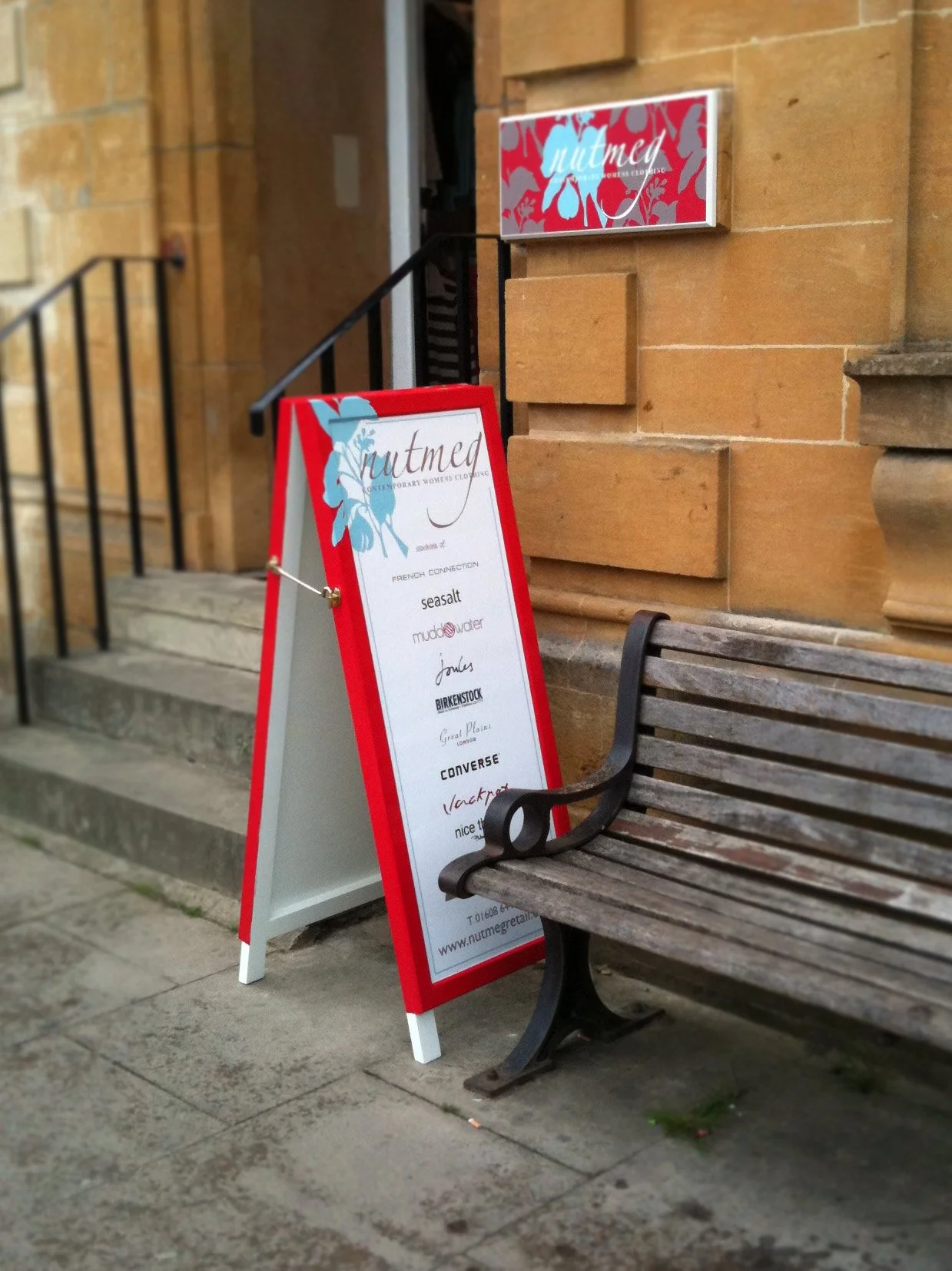 A sidewalk iron bench next to a red and white A-frame sign outside a building made of tan bricks. The sign displays various store brands and a website address.