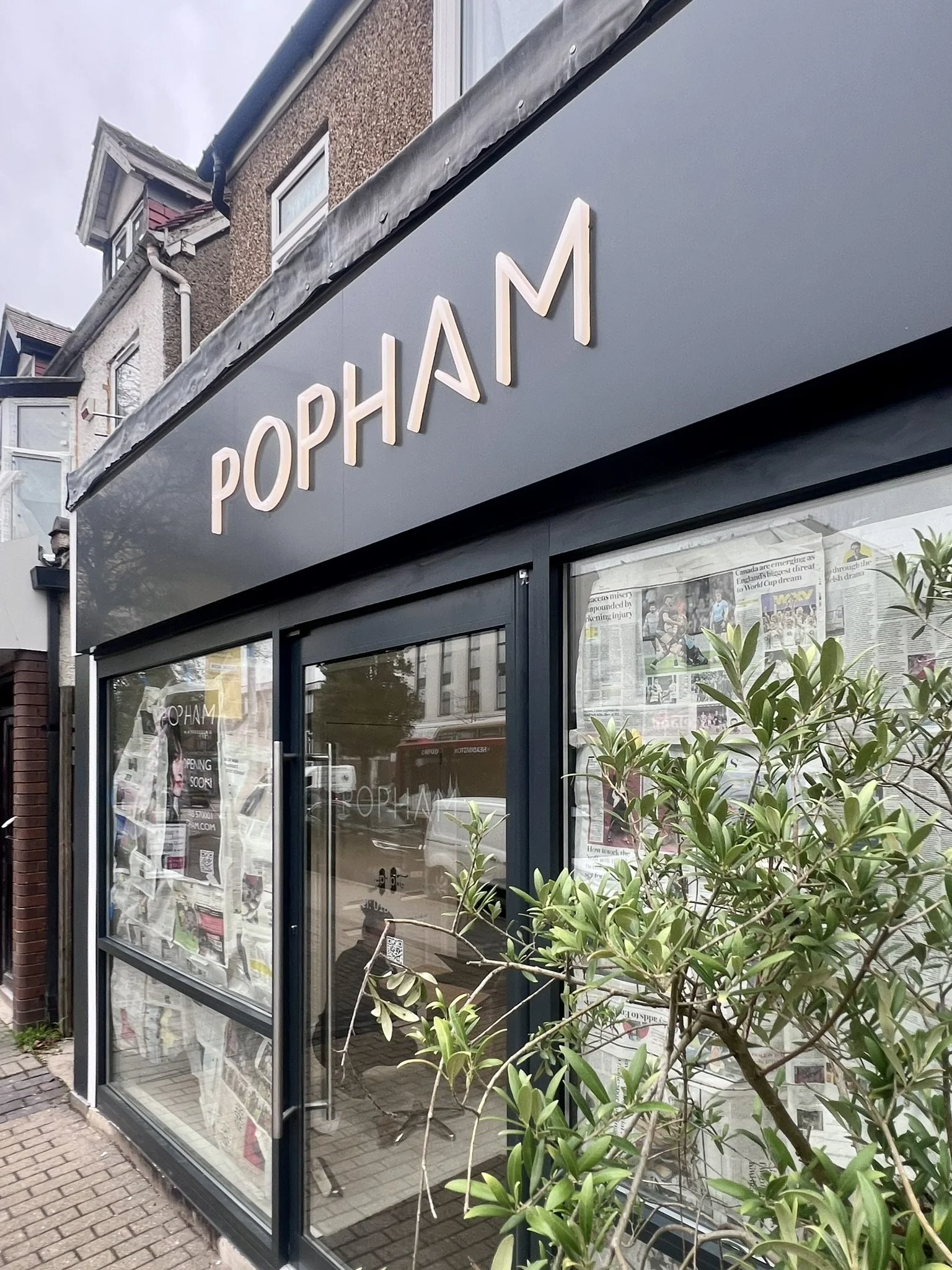 Exterior of a shop named POPHAM with large glass windows covered with newspaper and a small green bush in front.