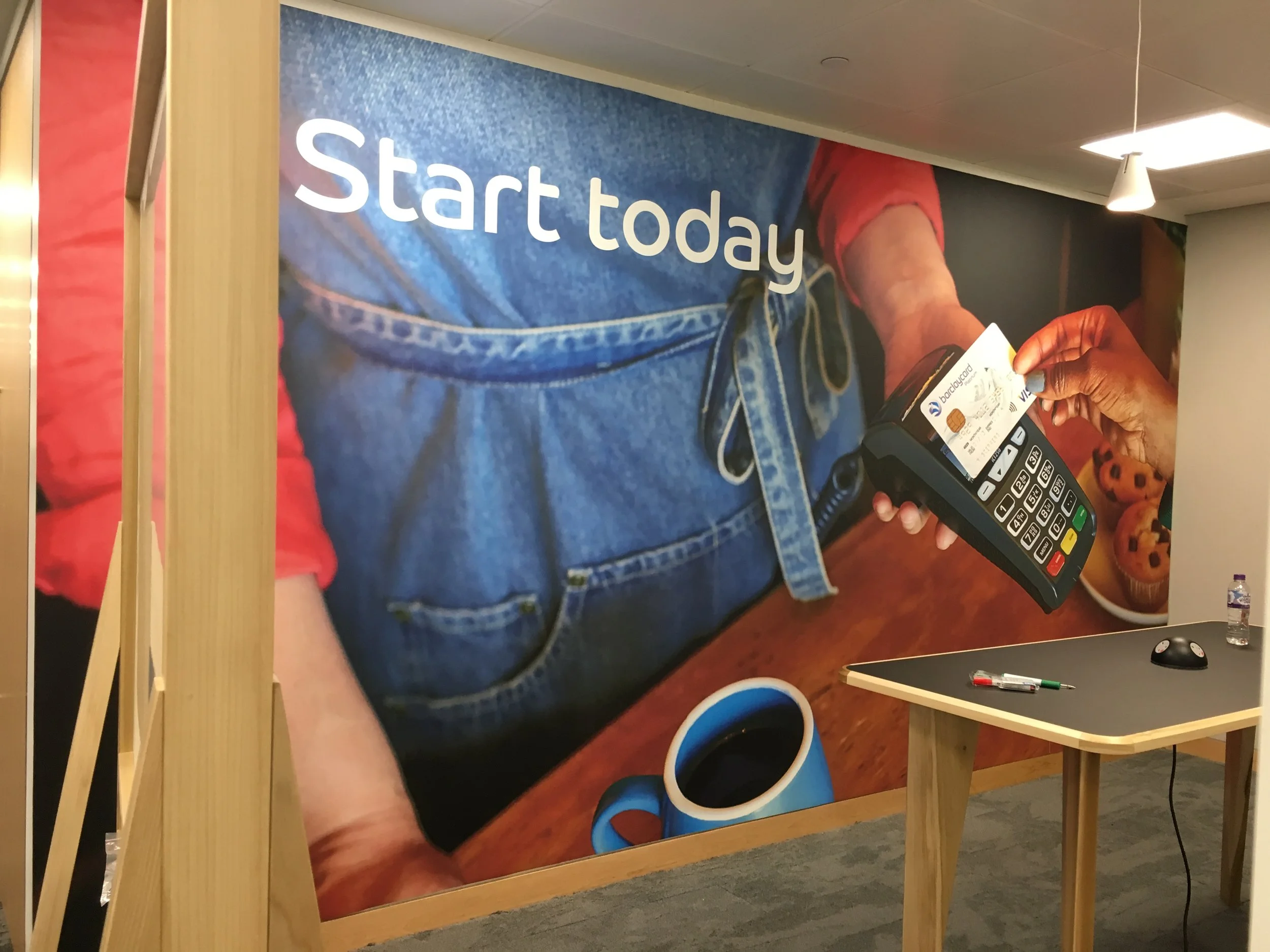 Large wall mural displaying a close-up of a person in a red shirt and blue apron using a point-of-sale terminal. The text 'Start today' is prominently displayed on the mural. A table in front holds a pen, a computer mouse, and a water bottle. A cup of coffee and some muffins are visible in the background in the mural.