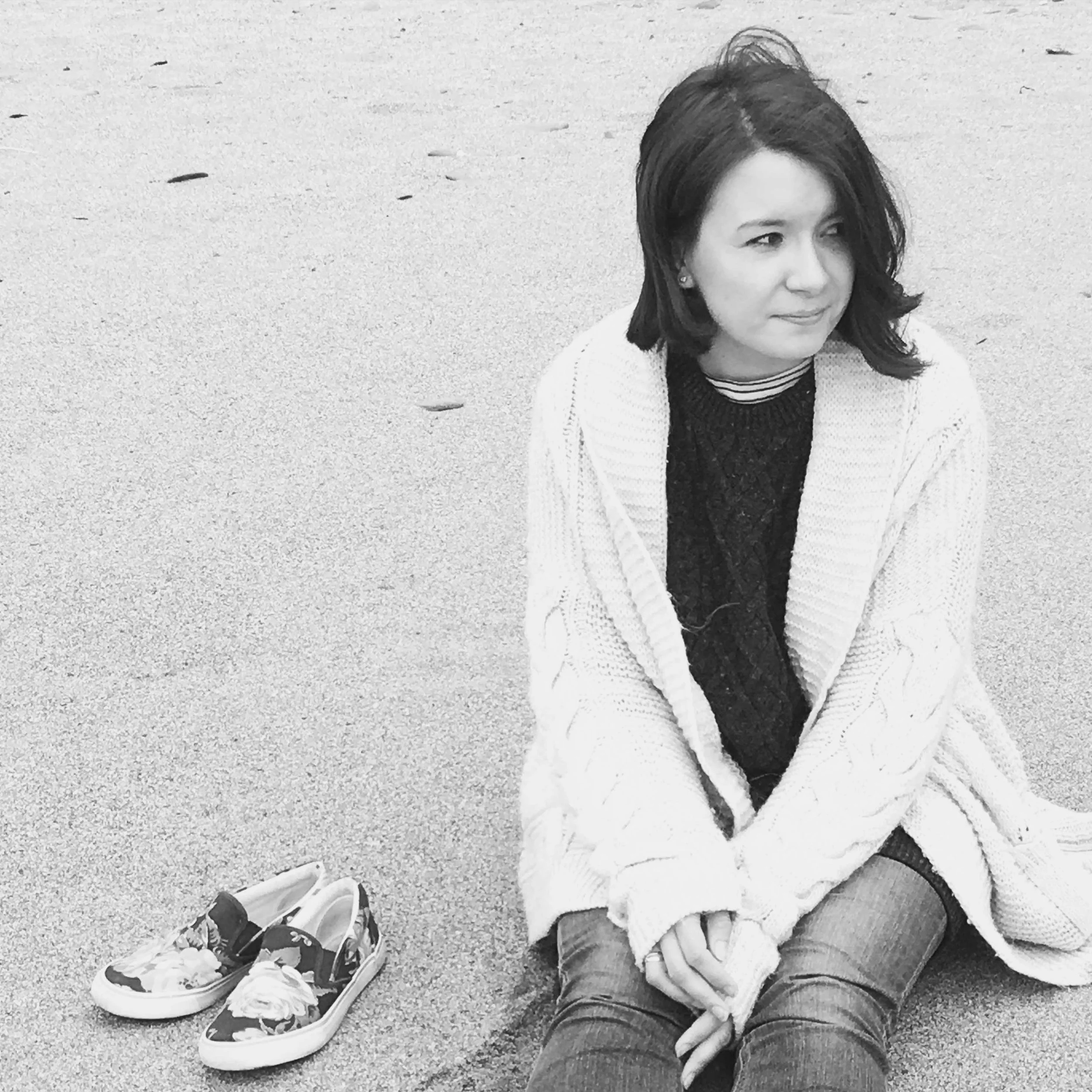 A woman sitting on a sandy surface with a pair of floral sneakers beside her, looking to her right with a slight smile.
