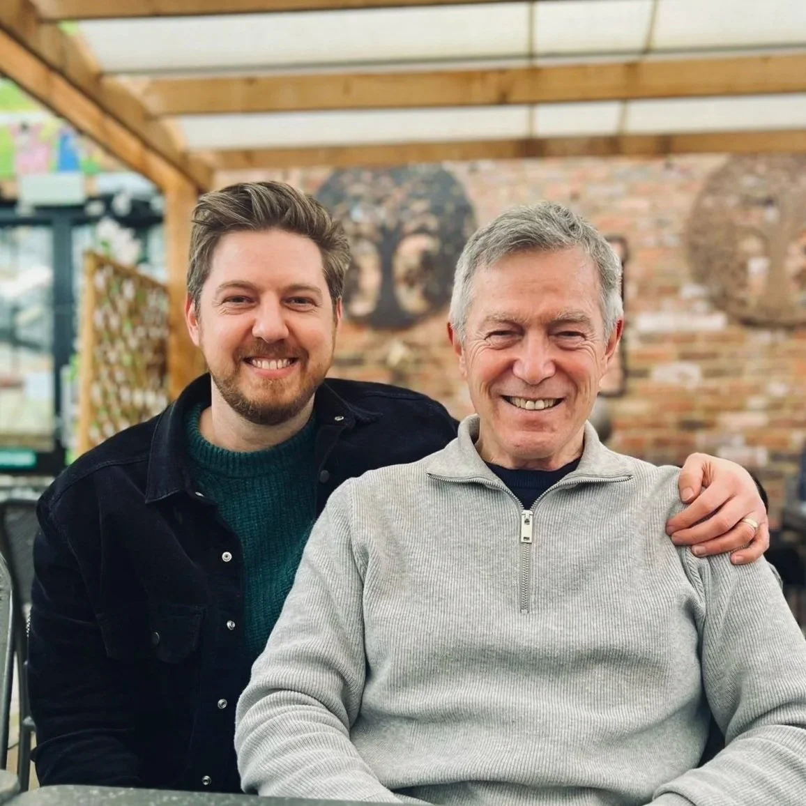 Two men smiling, sitting close together with one older man having his arm around the other in a cozy restaurant or cafe with brick walls and decorative artwork in the background.