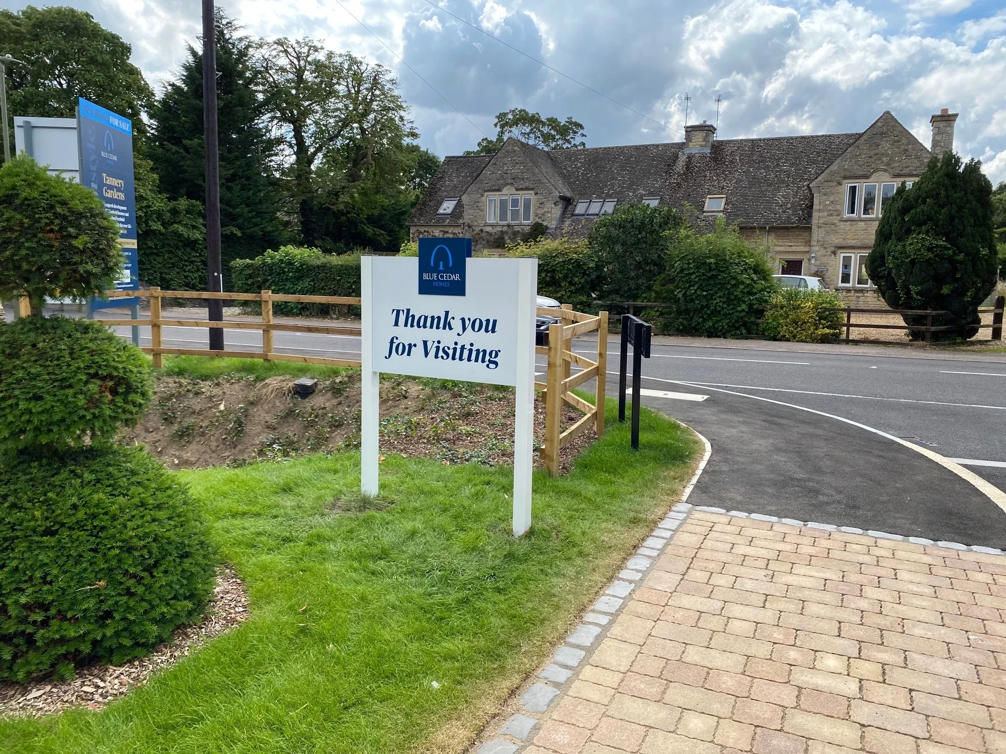 Sign that says 'Thank you for Visiting' in front of a stone house with a garden and parking lot. Blue Cedar Homes sign is visible.