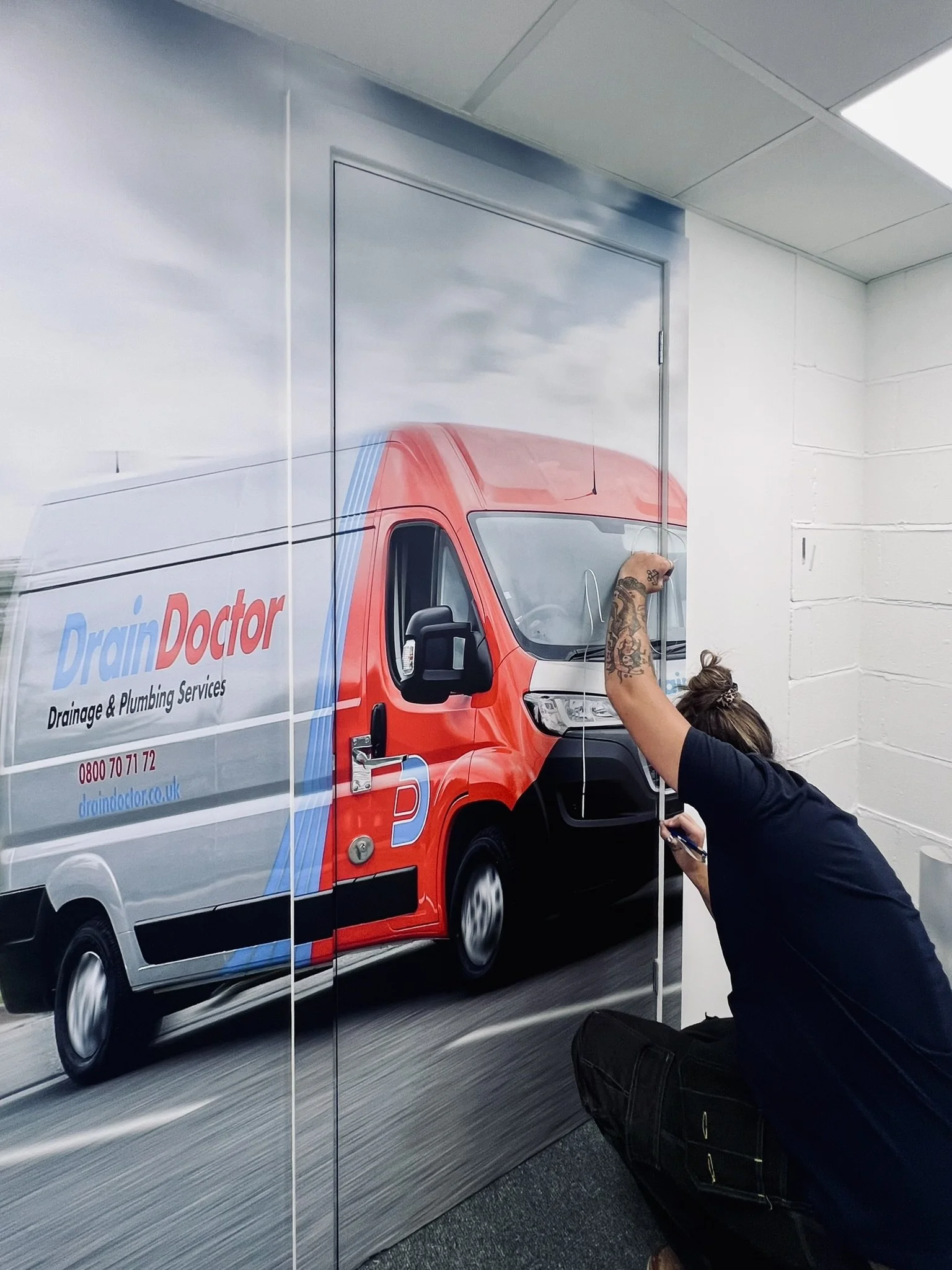 A person working on a wall mural of a delivery van with the text 'Drain Doctor Drainage & Plumbing Services'. The mural depicts a red and white van with the company's contact information.