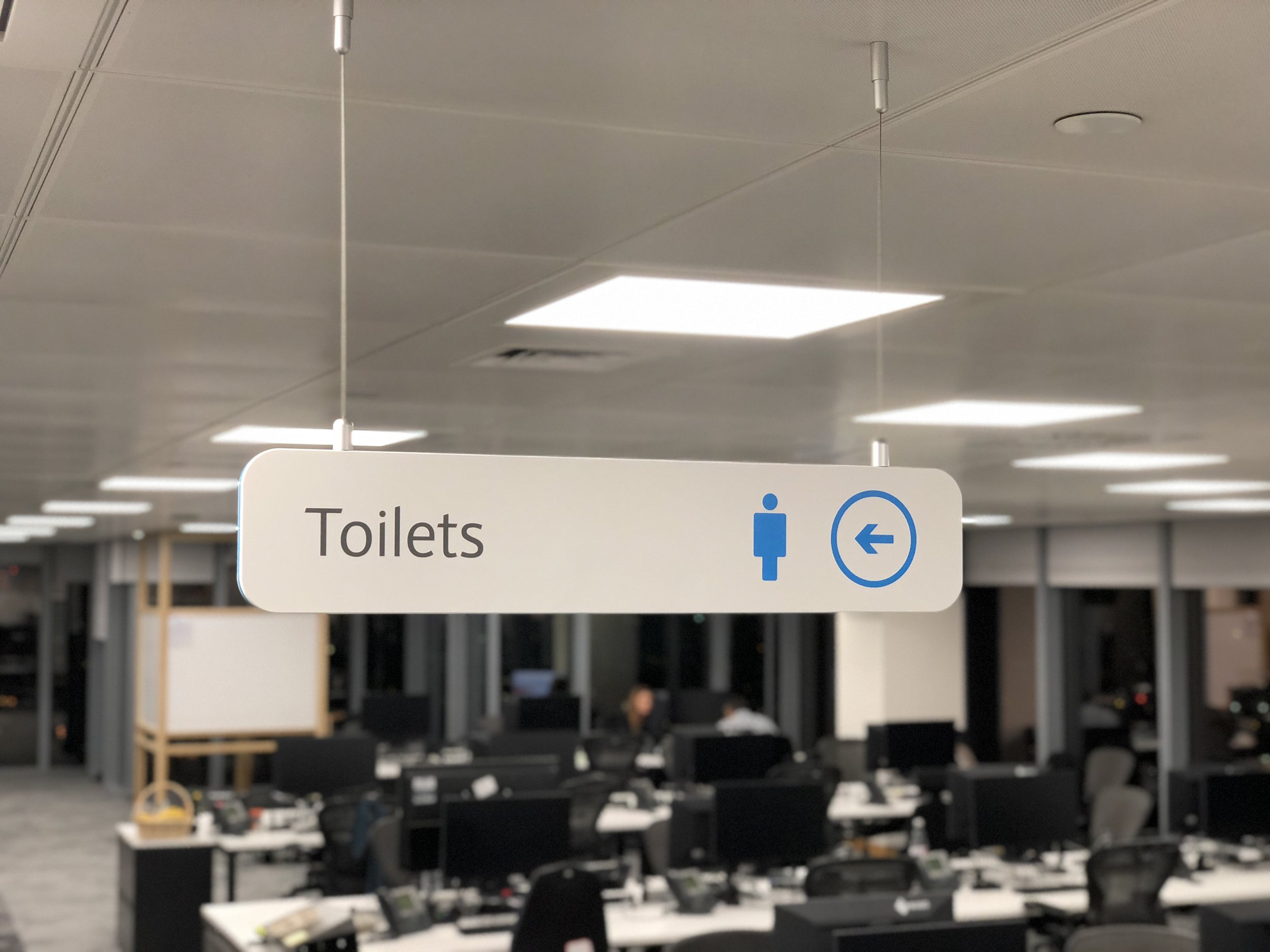 Hanging sign indicating directions to toilets in an office environment with desks and computers