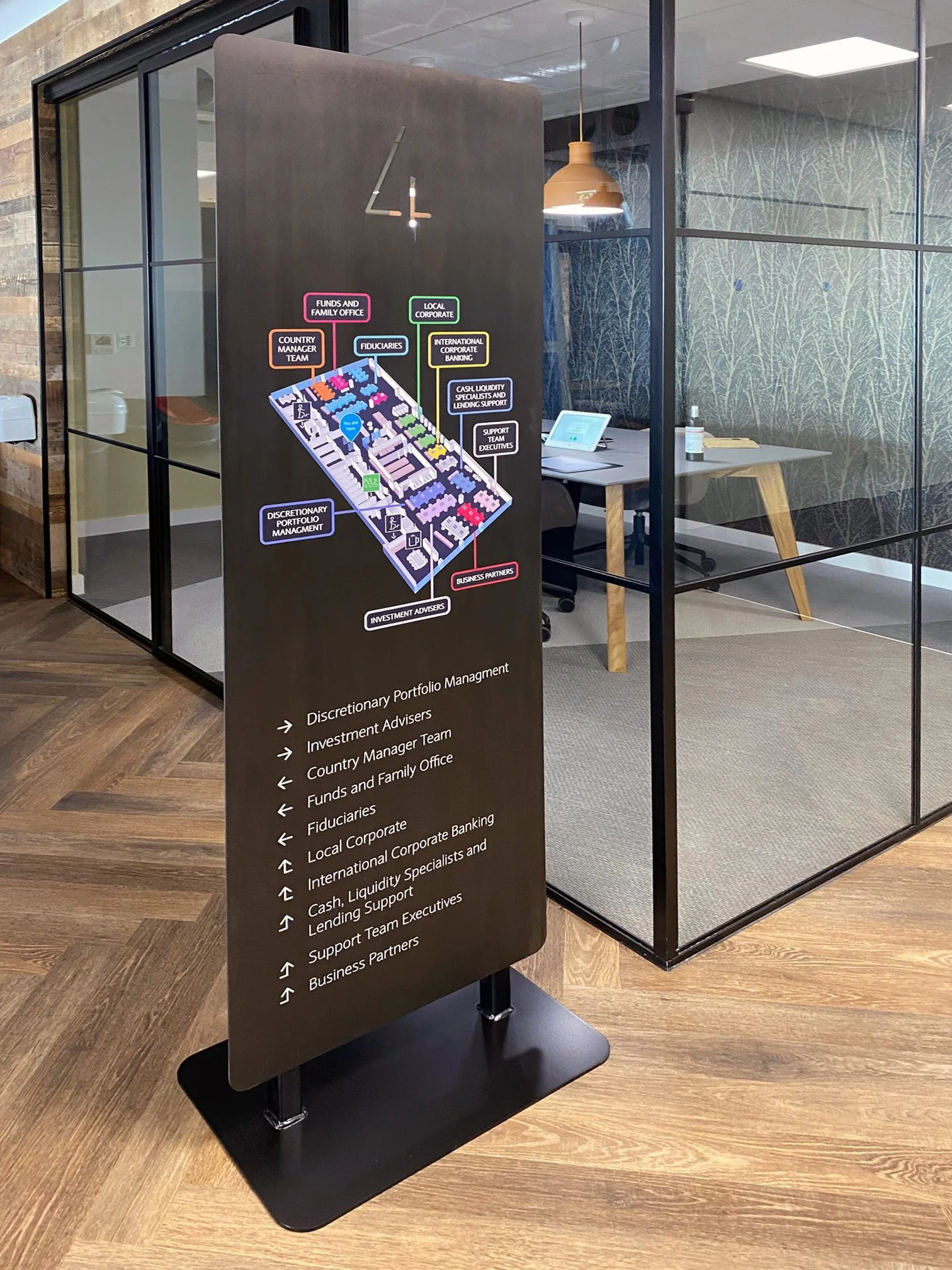Directory board with map and labels for various office departments including Portfolio Management, Investment Advisers, Country Manager Team, Funds and Family Office, Fiduciaries, Local Corporate, International Corporate Banking, Cash Liquidity Specialists and Lending Support, Support Team Executives, Business Partners, placed inside a modern office with glass walls, a desk with a tablet, and a hanging pendant light.