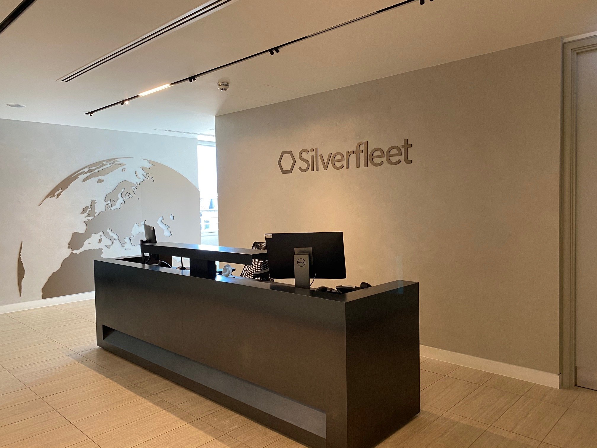 Office reception area with a black reception desk, two monitors, a wall sign reading 'Silverfleet,' and a partial world map mural on the wall to the left.