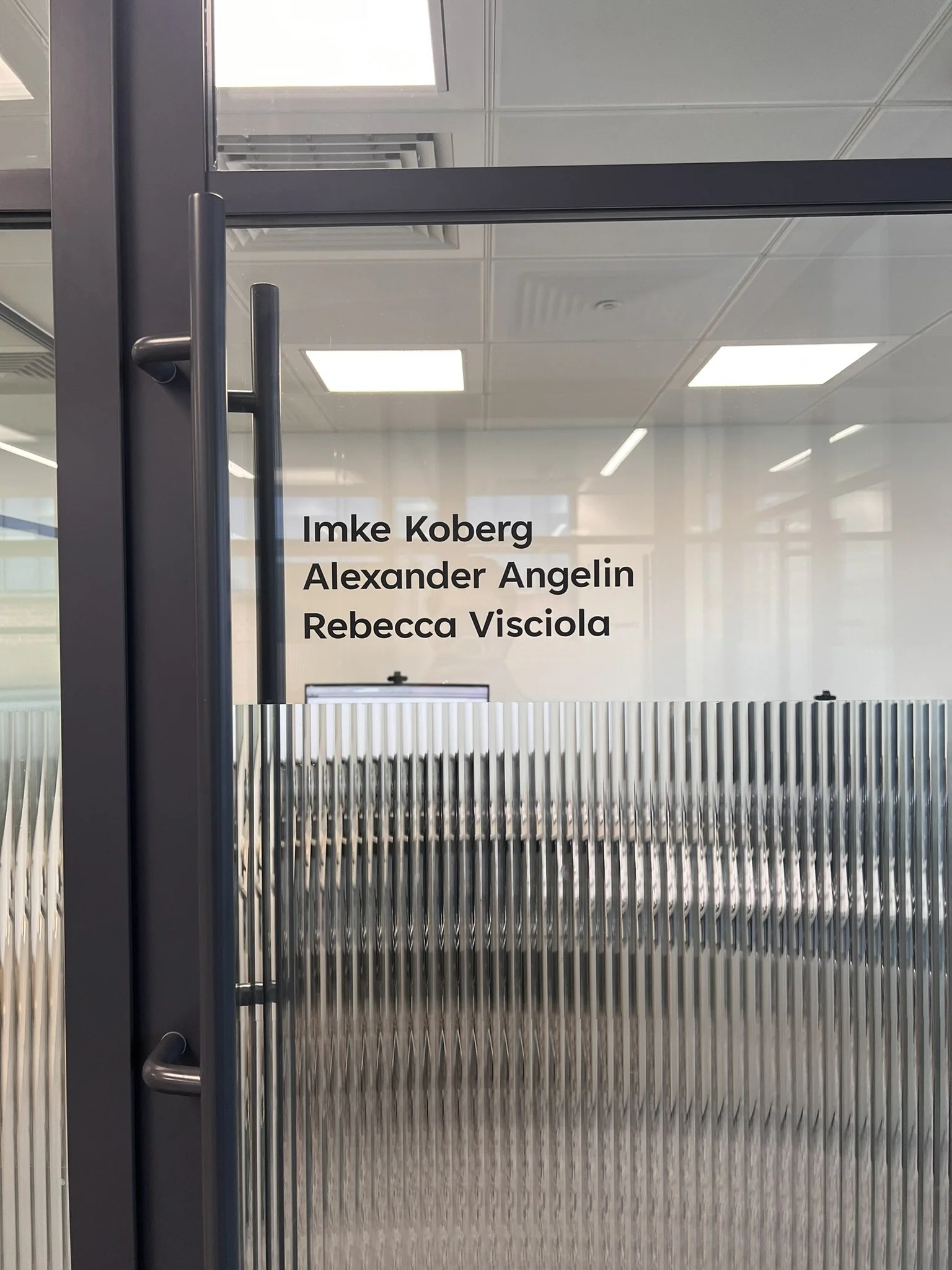 Names of three individuals: Imke Koberg, Alexander Angelin, and Rebecca Visciola are written on a glass door in an office setting.