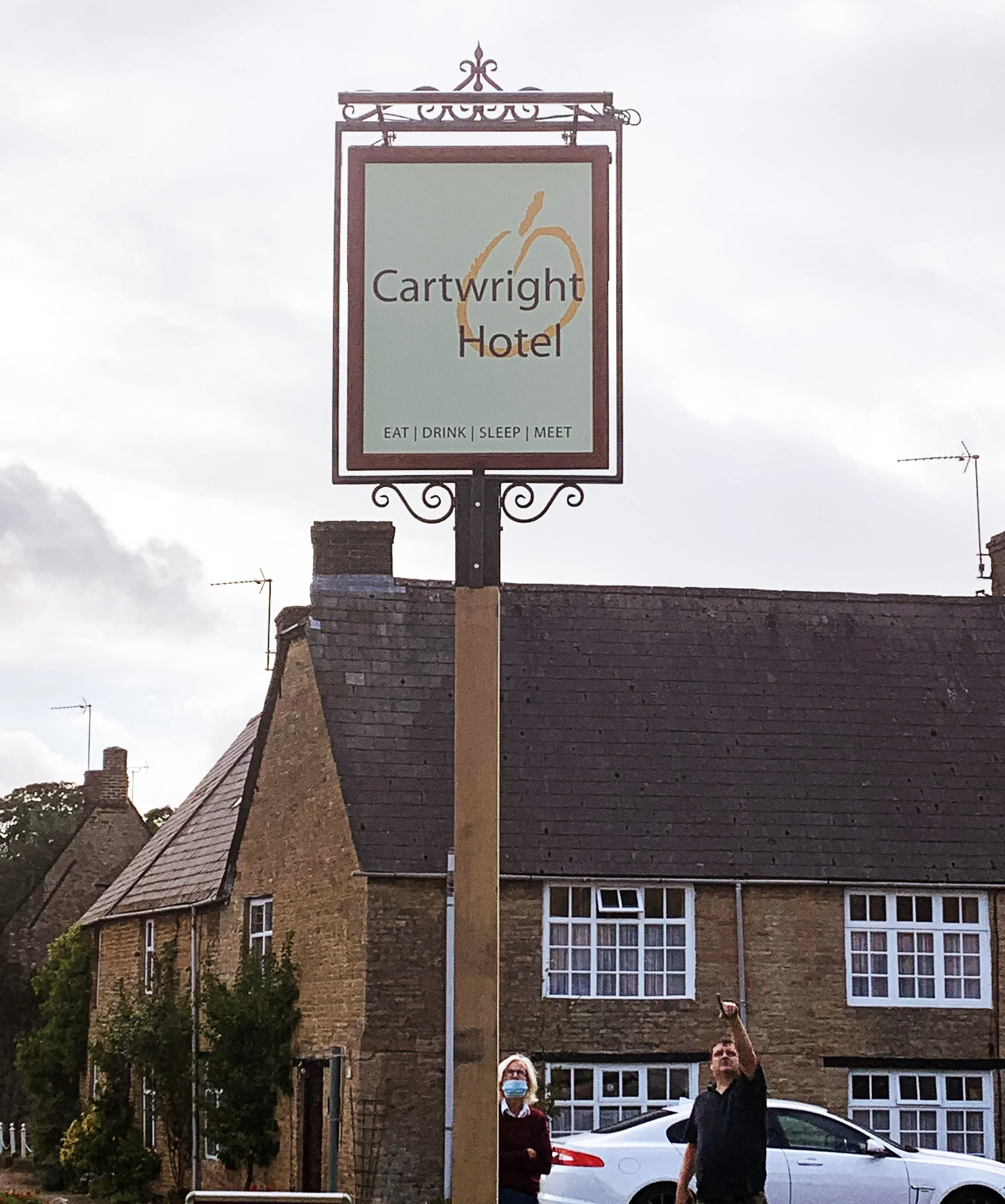 A sign for the Cartwright Hotel featuring a large apple graphic and the words 'Eat, Drink, Sleep, Meet' above a building with a sloped roof and several windows. Two people, one wearing a face mask, stand in front of the building under a cloudy sky.