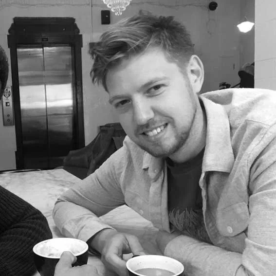 A young man with light hair and beard smiling at the camera while sitting at a table with two cups of coffee in a restaurant or cafe.