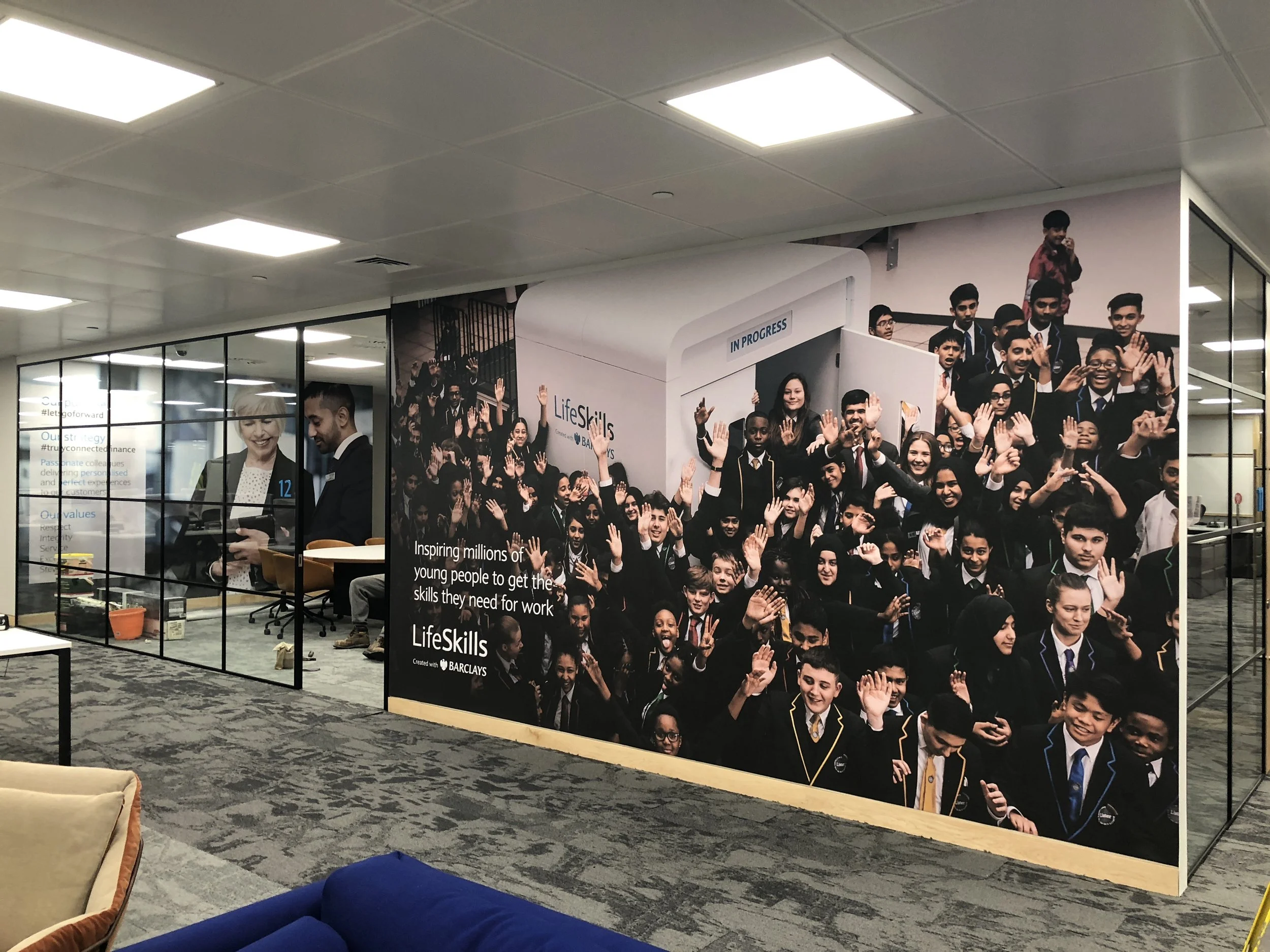Office space with a glass enclosed meeting room on the left, featuring a man and woman working on a laptop. Outside the room, there's a large wall mural of a diverse group of young students in school uniforms, smiling and waving, with a caption about inspiring young people to acquire work skills.