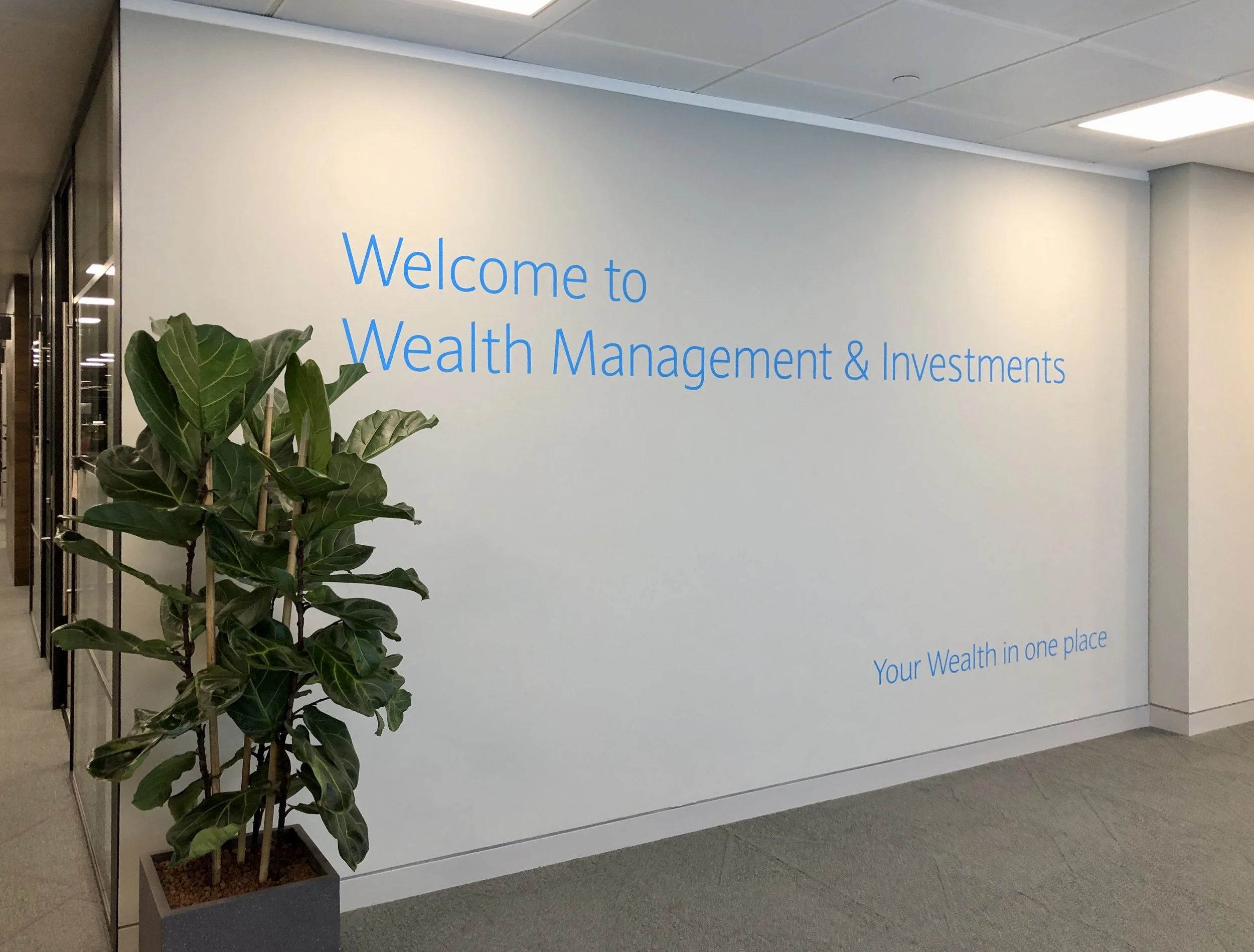 Office wall with welcome message for Wealth Management & Investments, rounded corner, potted plant on the left, phrase 'Your Wealth in one place' at the bottom right.
