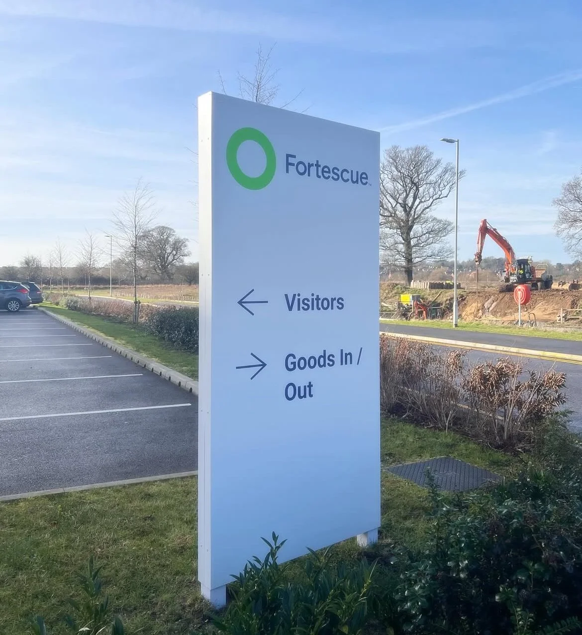 Sign for Fortescape with directions for visitors to the left and goods in/out to the right at a parking lot outside, with some trees, a construction site, and a clear blue sky in the background.