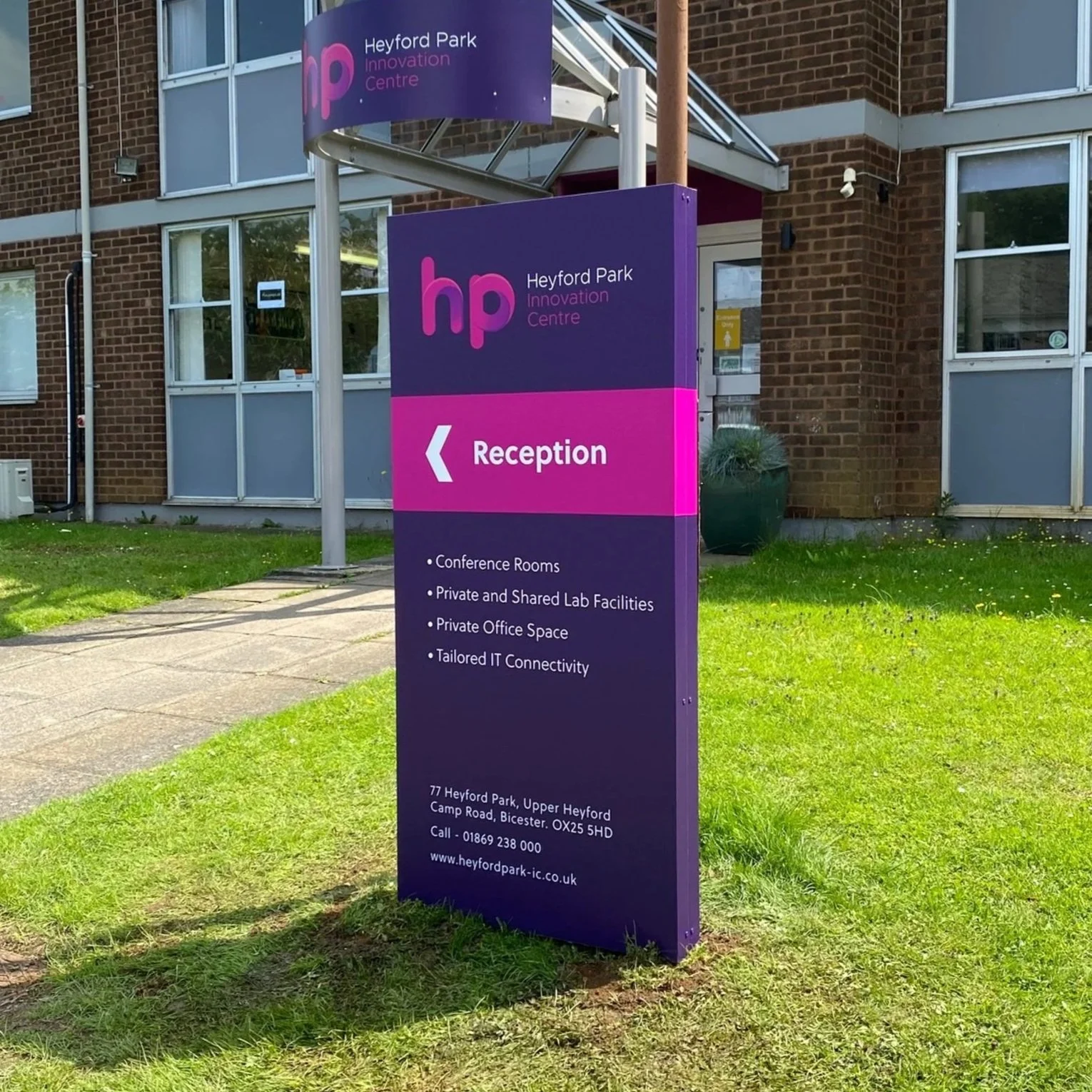 Sign outside Heyford Park Innovation Centre indicating the location of the reception with an arrow and listing facilities like conference rooms, lab facilities, private office space, and IT connectivity. The sign has purple and pink colors and is situated on a grassy area near a brick building.