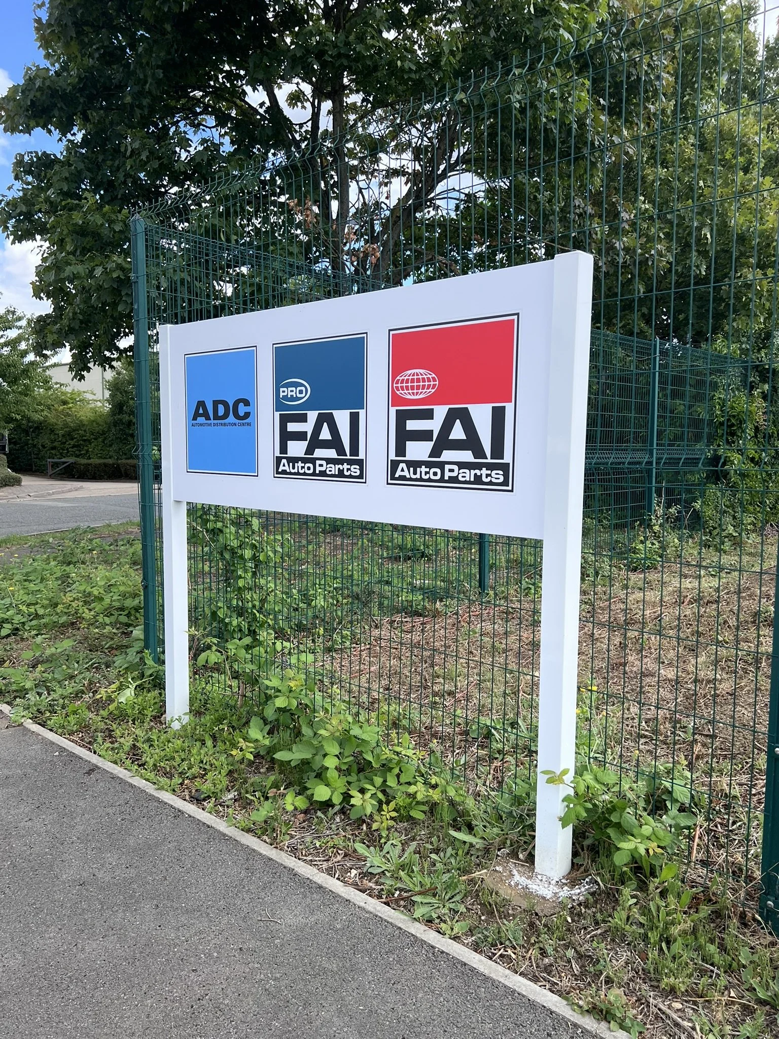 Sign with three logos for automotive parts stores: ADC, FAI PRO, and FAI Auto Parts, placed next to a green wire fence and some greenery.