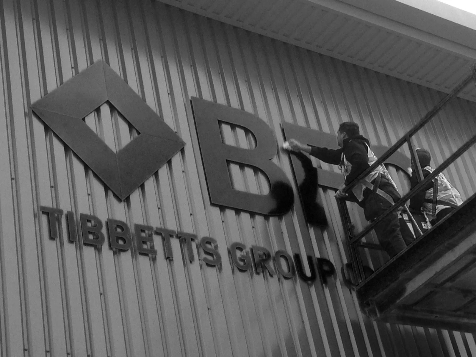 Two workers on a scaffold installing or cleaning a large sign on the exterior wall of a building. The sign reads 'DRE' in large letters, with smaller text below that says 'TIBBETTS GROUP.'