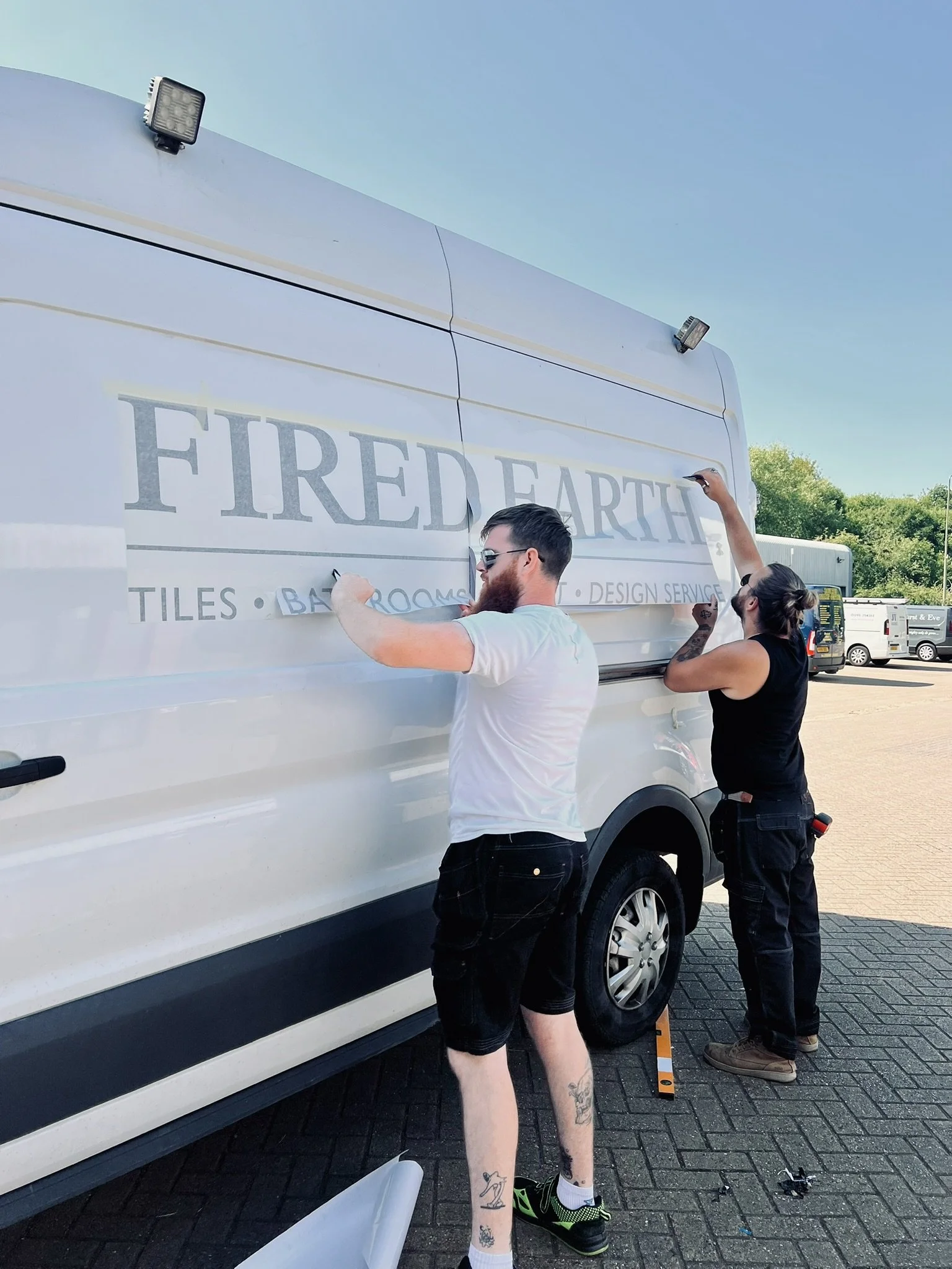 Two people are applying a vinyl decal to the side of a white van. The decal reads 'FIRED EARTH' and mentions tiles, bathrooms, and design services. One person is wearing a white t-shirt and shorts, while the other is in a black t-shirt and pants.