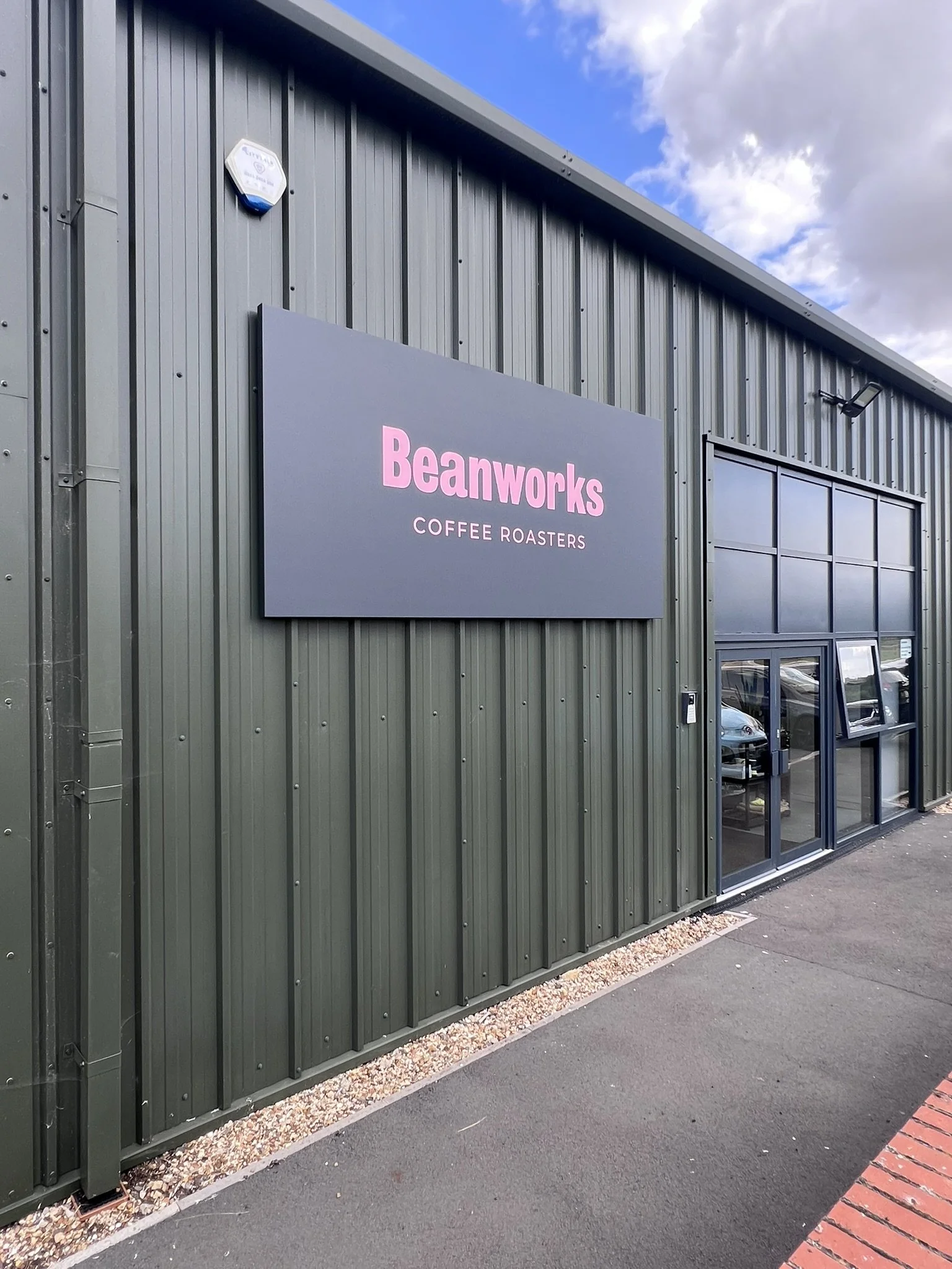 Exterior of Beanworks Coffee Roasters building with a sign on a dark green metal wall, and a glass garage door reflecting parked cars and cloudy sky.