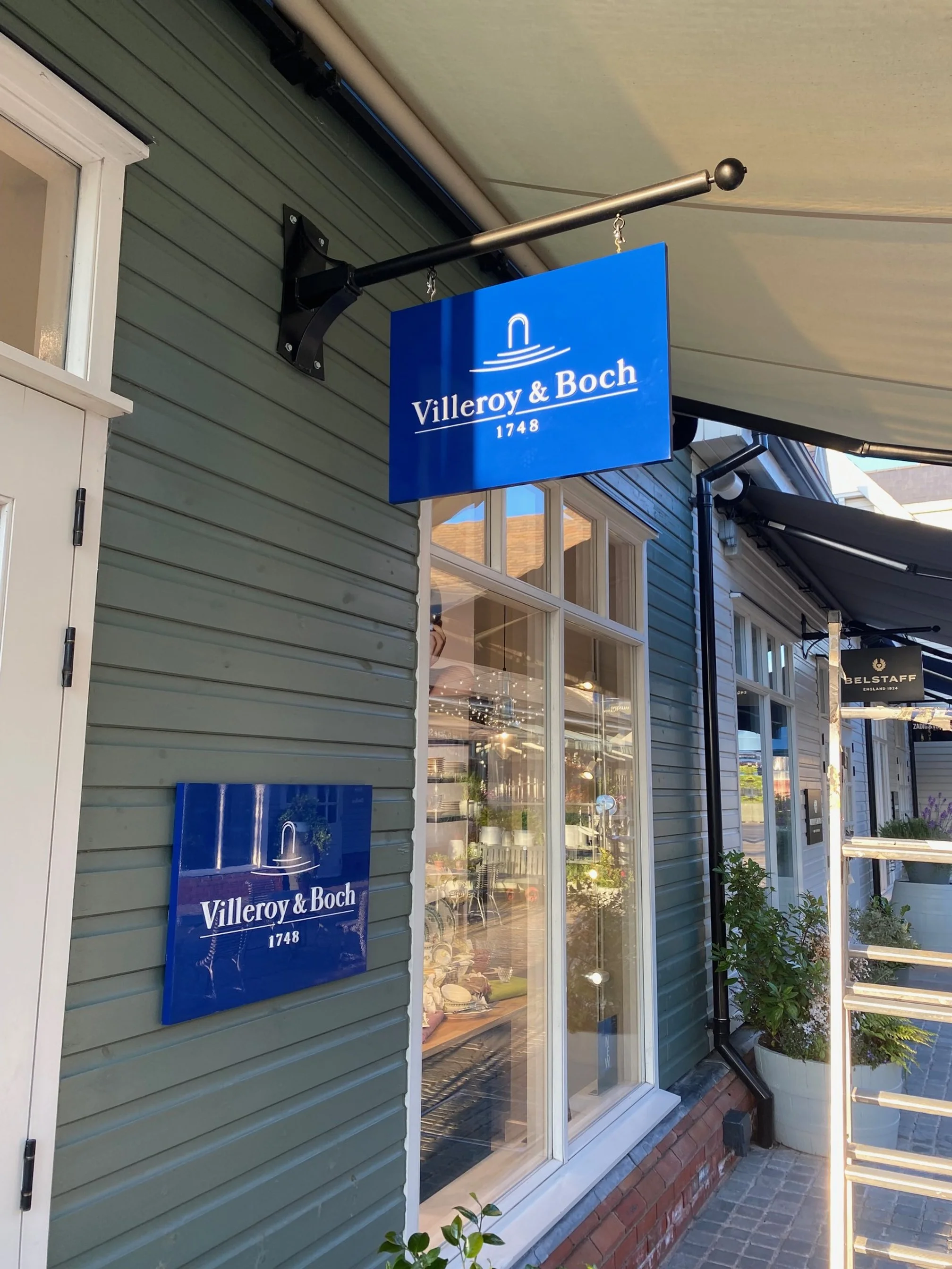 Storefront with blue signs for Villeroy & Boch, number 1748, and a glimpse of the shop interior through large windows, with plants outside.