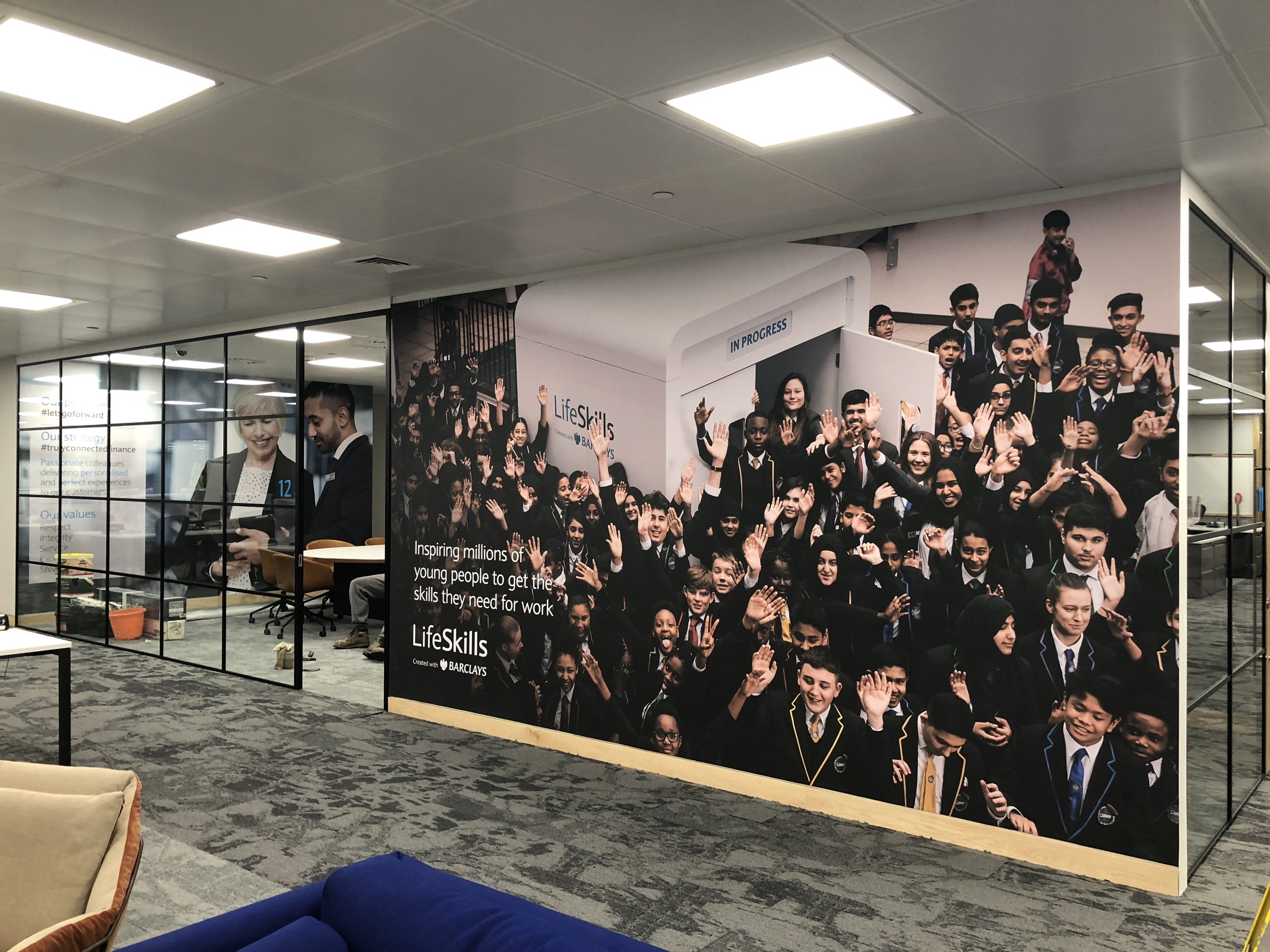 Large wall mural featuring a diverse group of school children and teenagers waving and smiling, some in uniforms, in front of a sign that says 'IN PROGRESS'. To the left, an office space with two men working at a desk behind a glass partition.