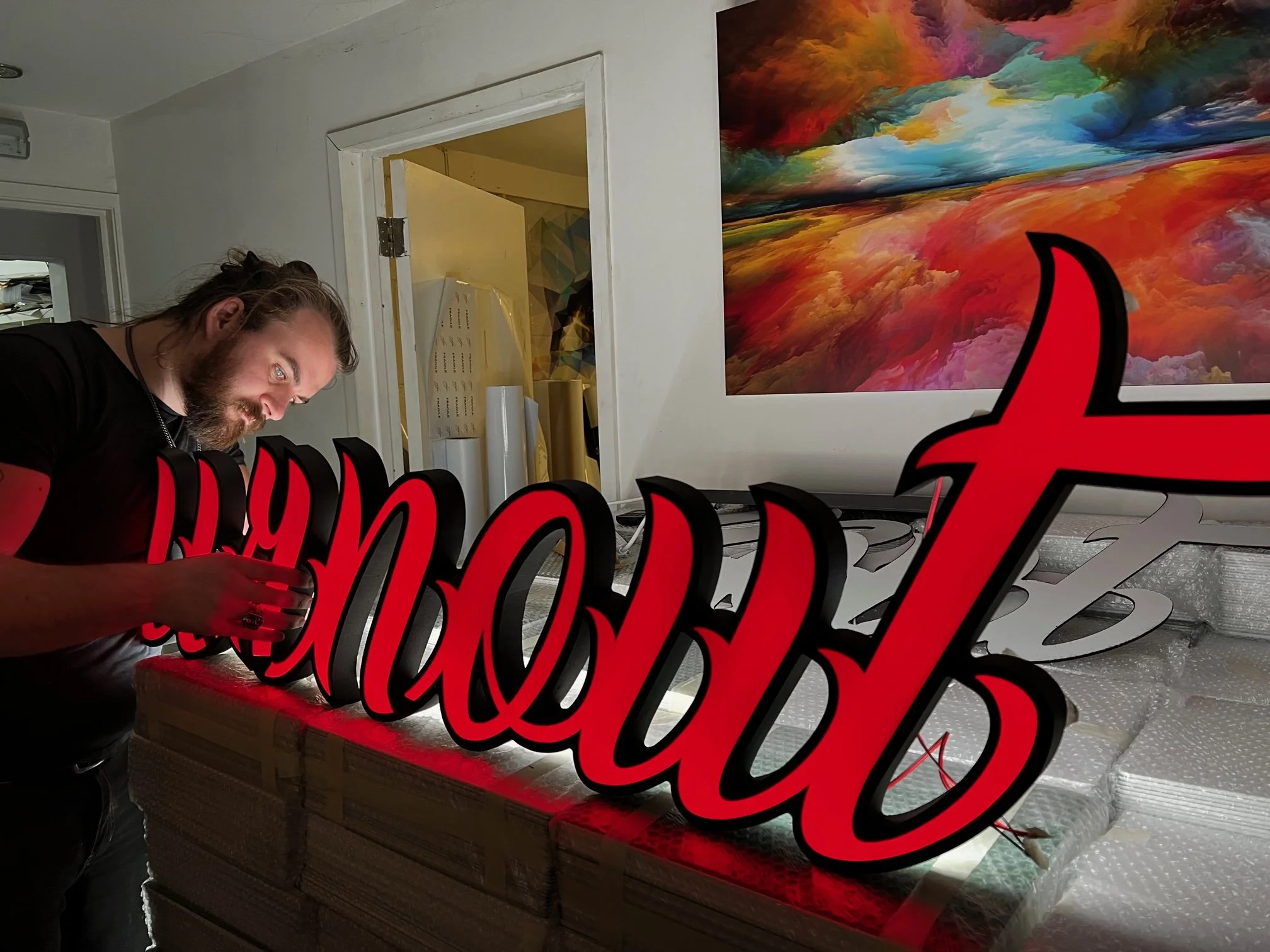 A man with long hair and a beard installing a red and black illuminated sign that says 'love' on a table covered with bubble wrap in an art studio or workshop, with a colorful abstract painting on the wall behind him.