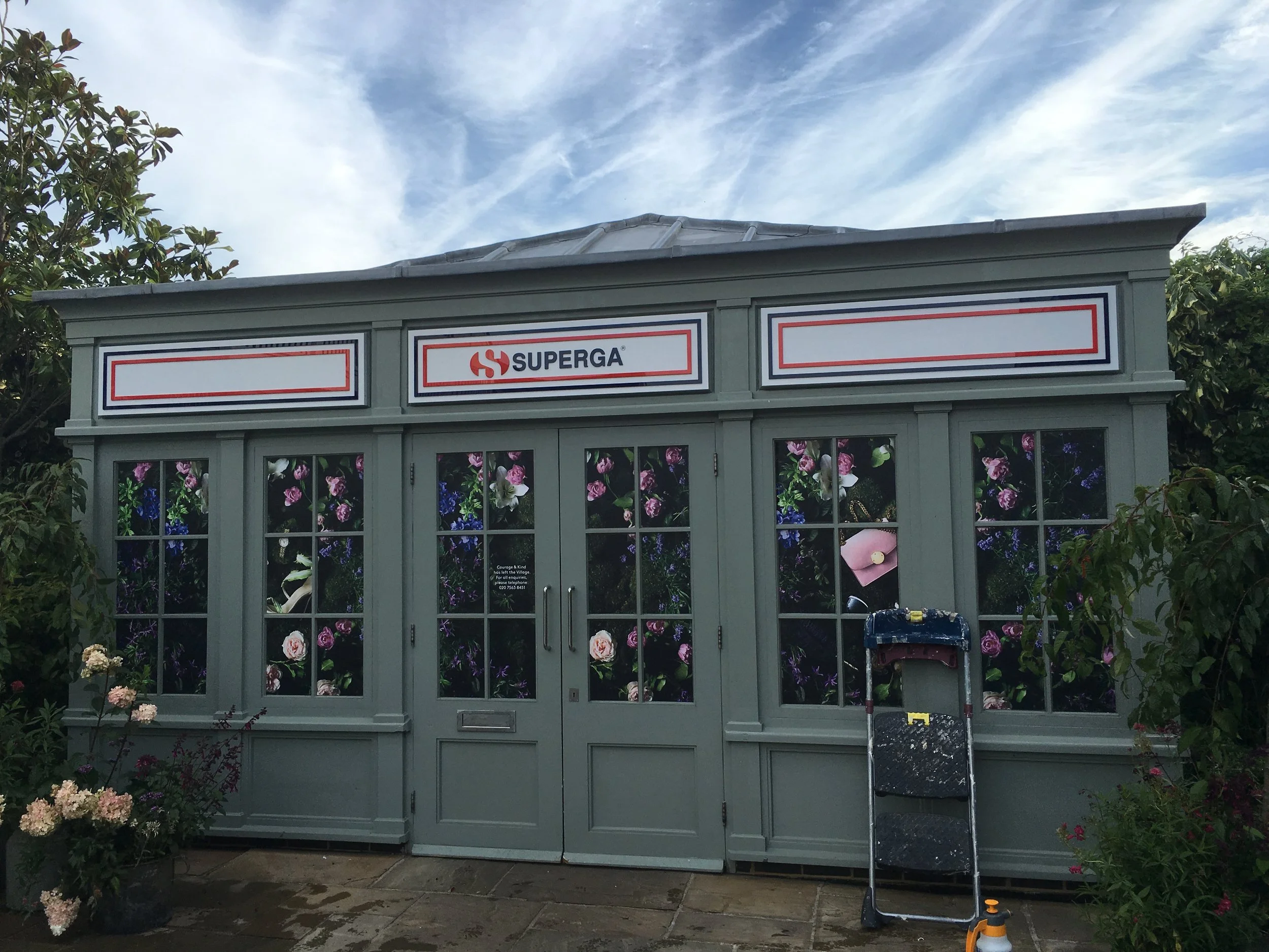 Gray garden shed with large windows displaying flower images, a ladder leaning against it, and a sign with the Superga logo on top, under a partly cloudy sky.