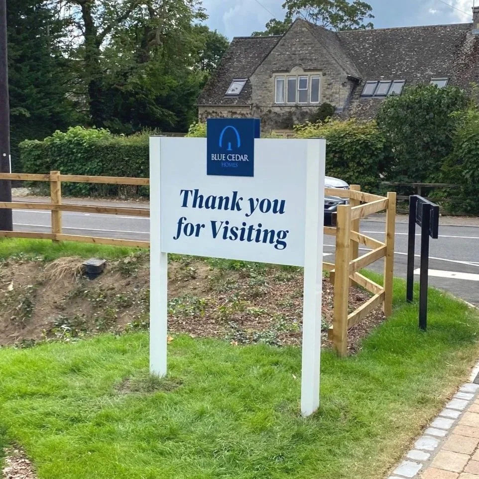A sign on a grassy area that says, "Thank you for Visiting" with a logo for Blue Cedar Homes, next to a sidewalk and a wooden fence, with houses and trees in the background.