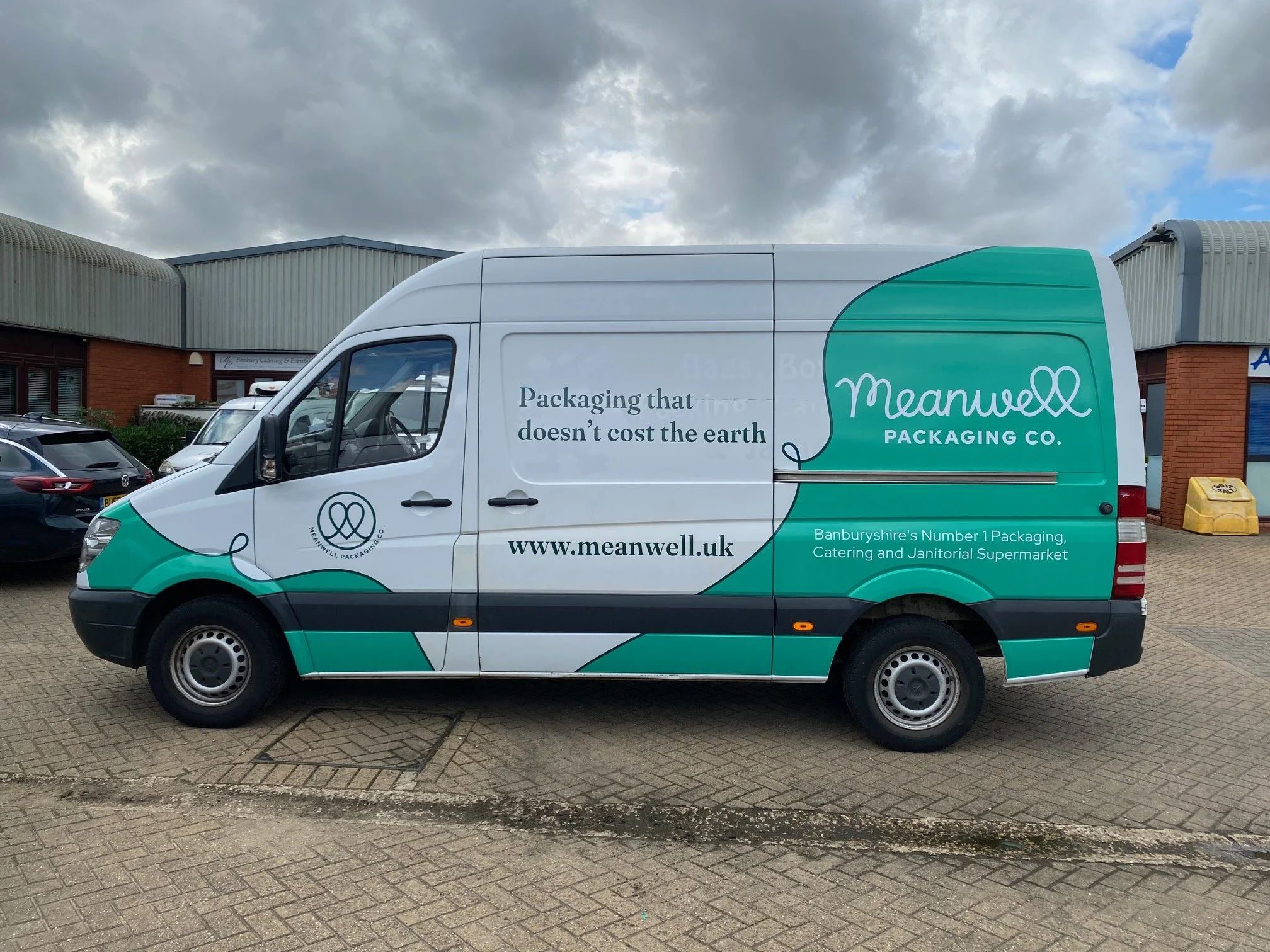 A delivery van with branding for Meanwell Packaging Co., parked outside a commercial building. The van has green and white graphics, with text promoting eco-friendly packaging and the company's website.