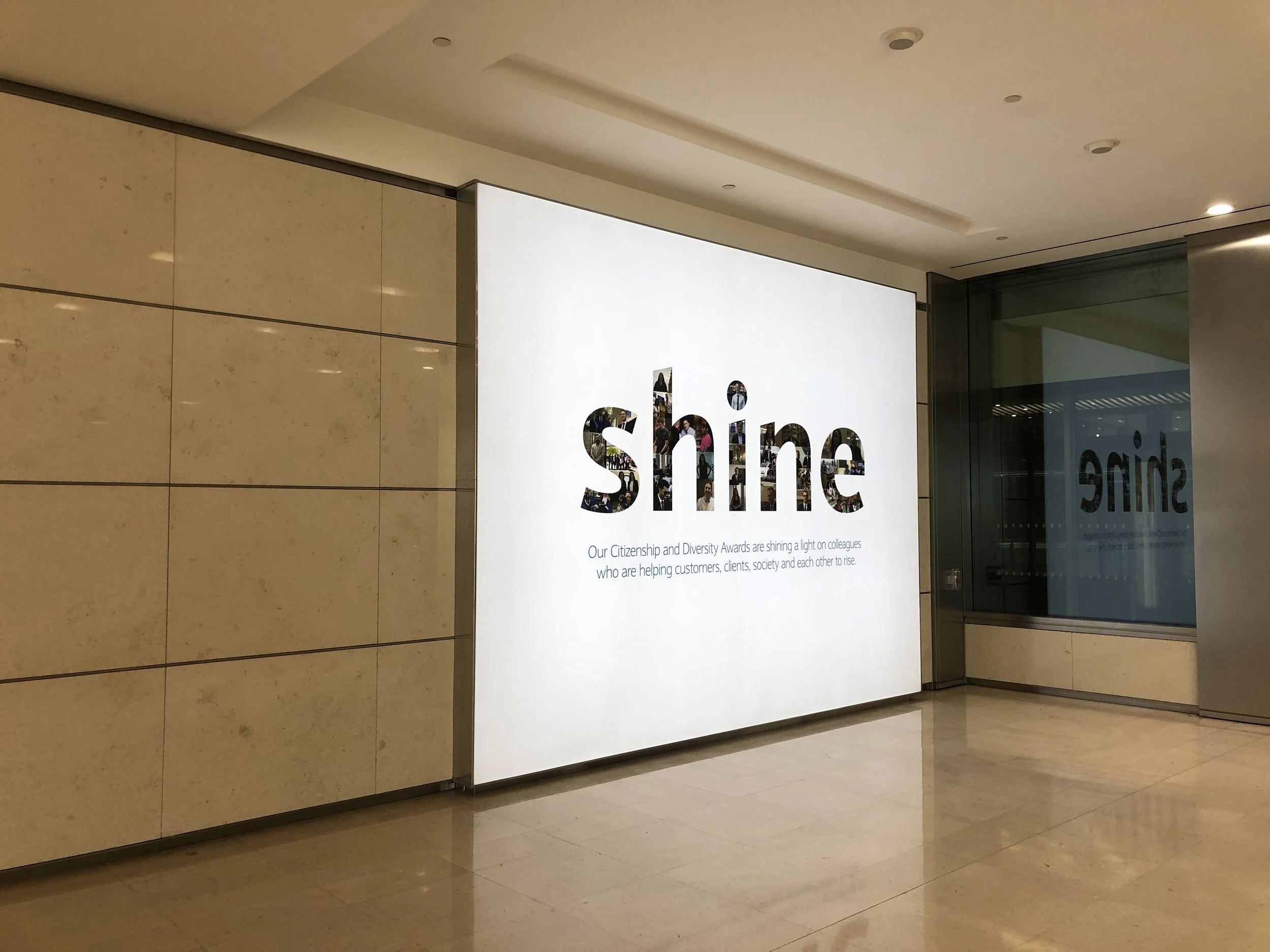 A lit up sign with the word 'Shine' on a white background, with smaller text below it about citizenship and diversity awards, inside a building with beige tiled walls and a large window reflecting the sign.