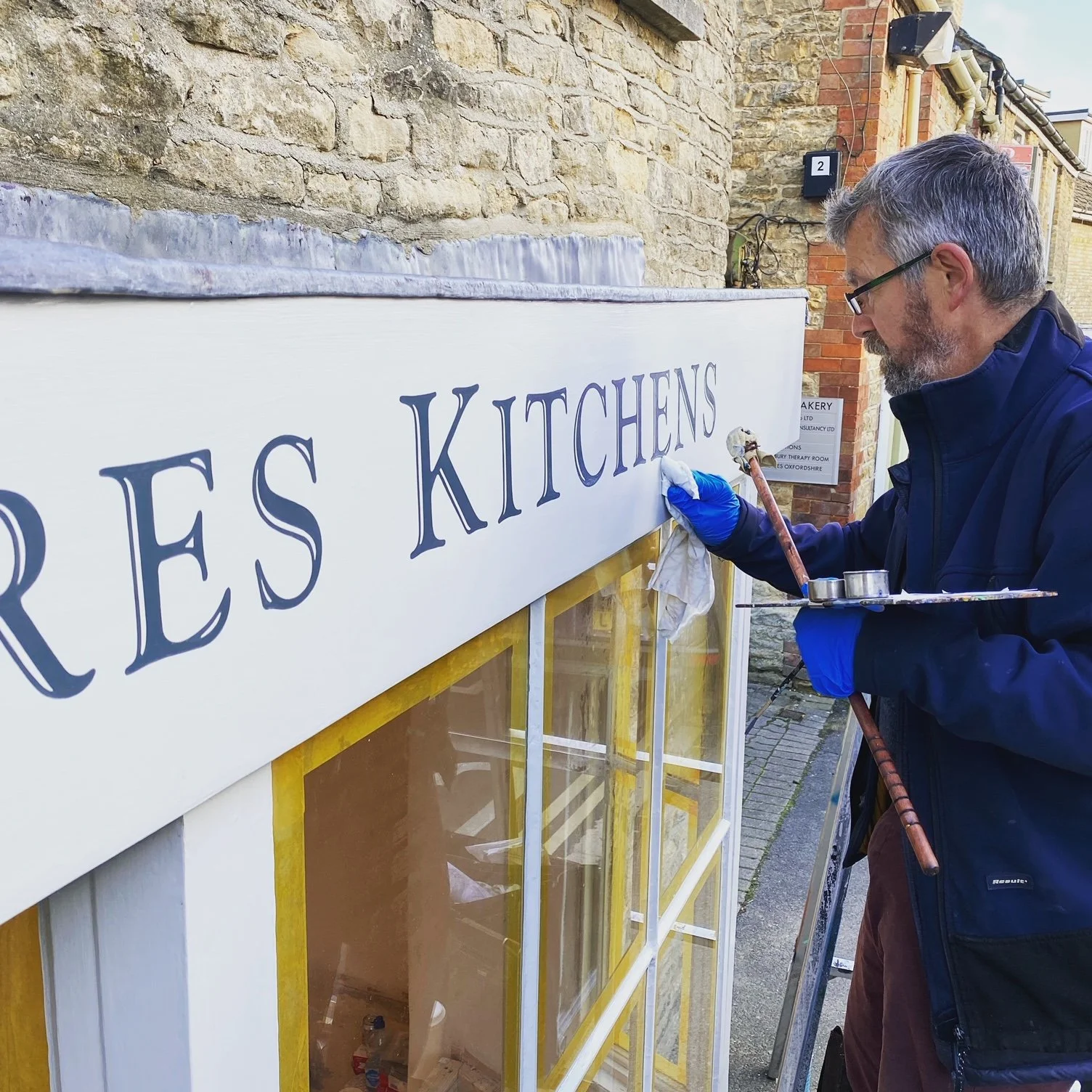 A man wearing glasses, a navy jacket, and blue gloves is painting the letters on a white sign that says 'GRORES KITCHENS' outside a building with brick and stone walls.