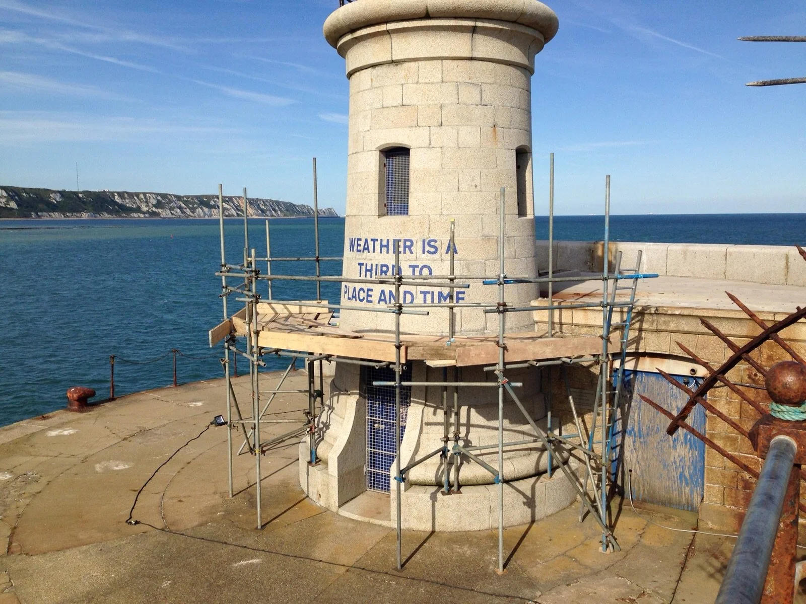 A lighthouse on a pier by the ocean with scaffolding around its base, and a partially visible sign that reads 'Weather is a third to place and time.'