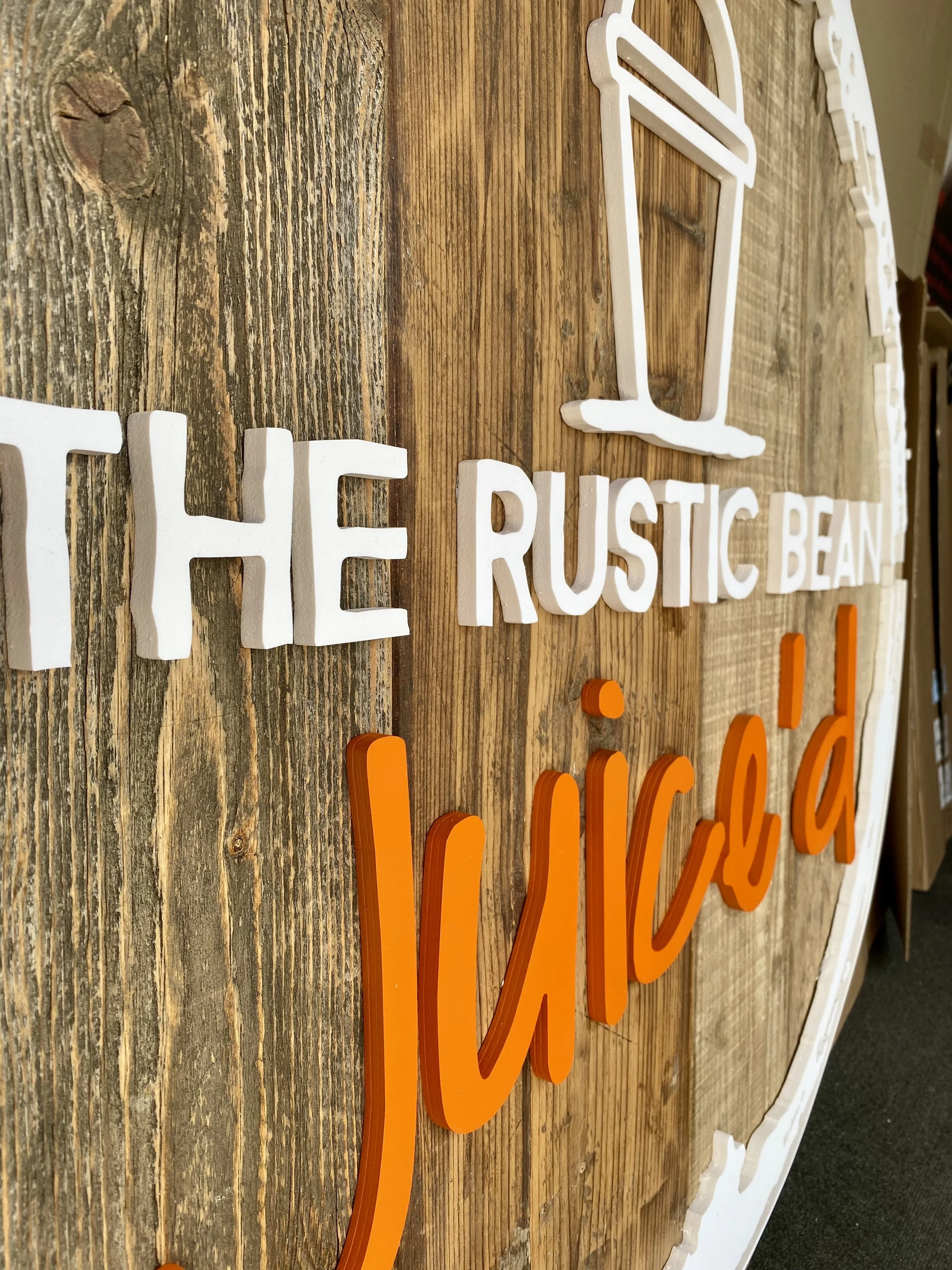 Wooden sign with white and orange 3D lettering that reads "The Rustic Bean Jukebox" and features a white outline of a jukebox.